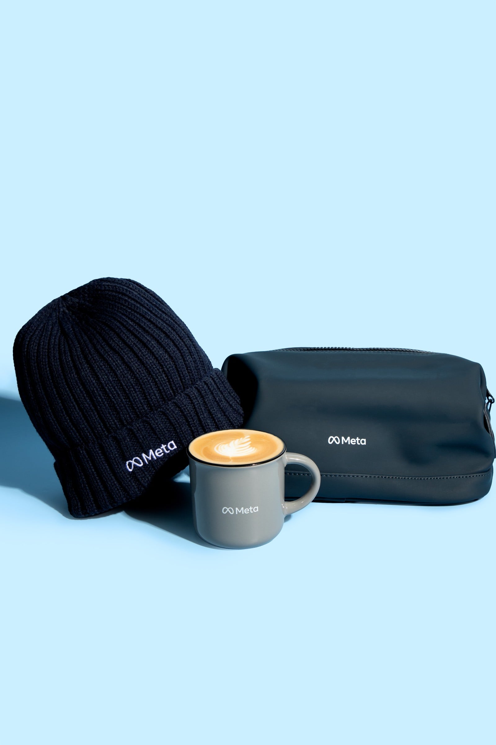 Meta branded navy beanie, coffee cup and pouch on a pale blue background.