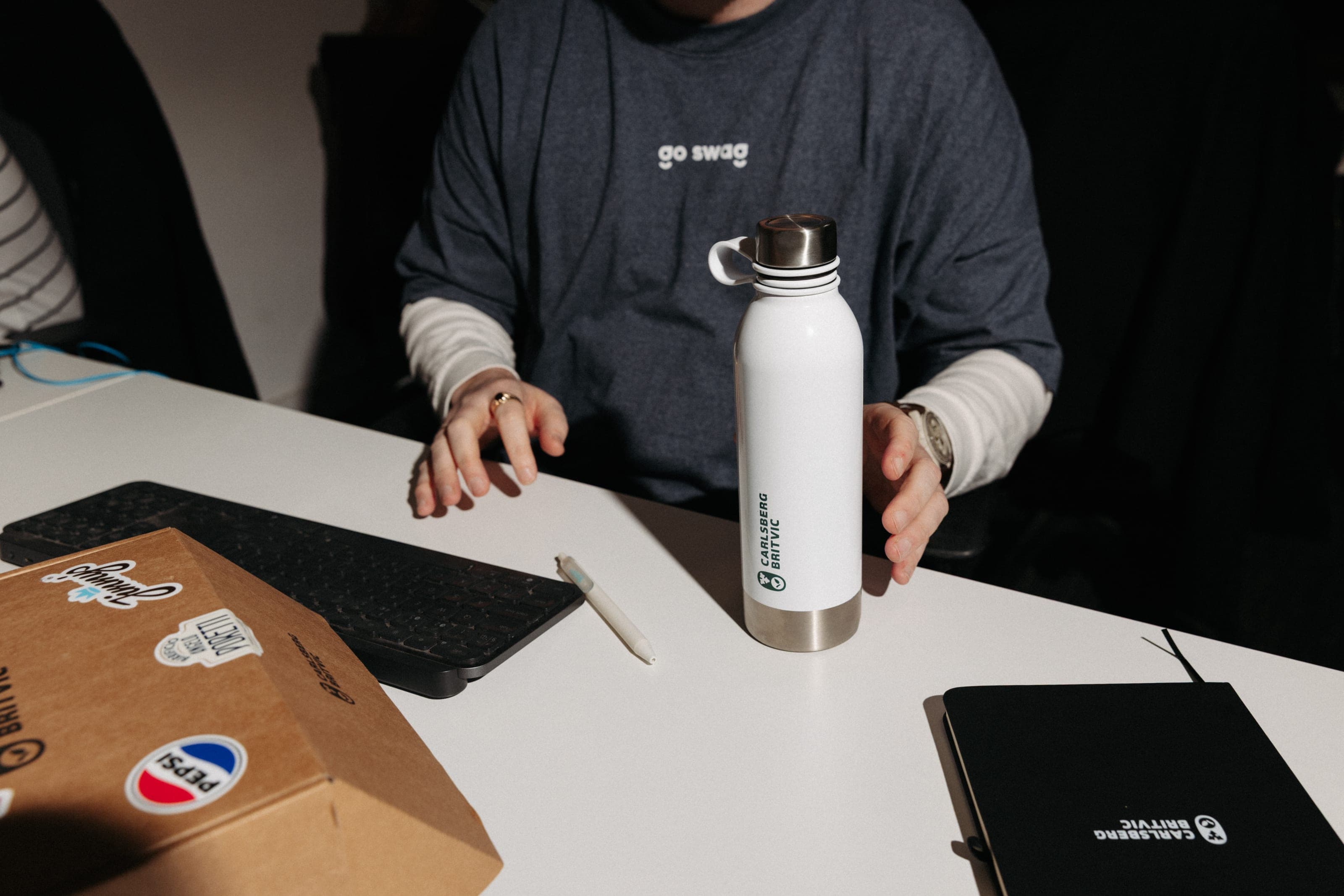A white Carlsberg Britvic bottle on a desk beside a stickered kraft box and a black Carlsberg Britvic notebook