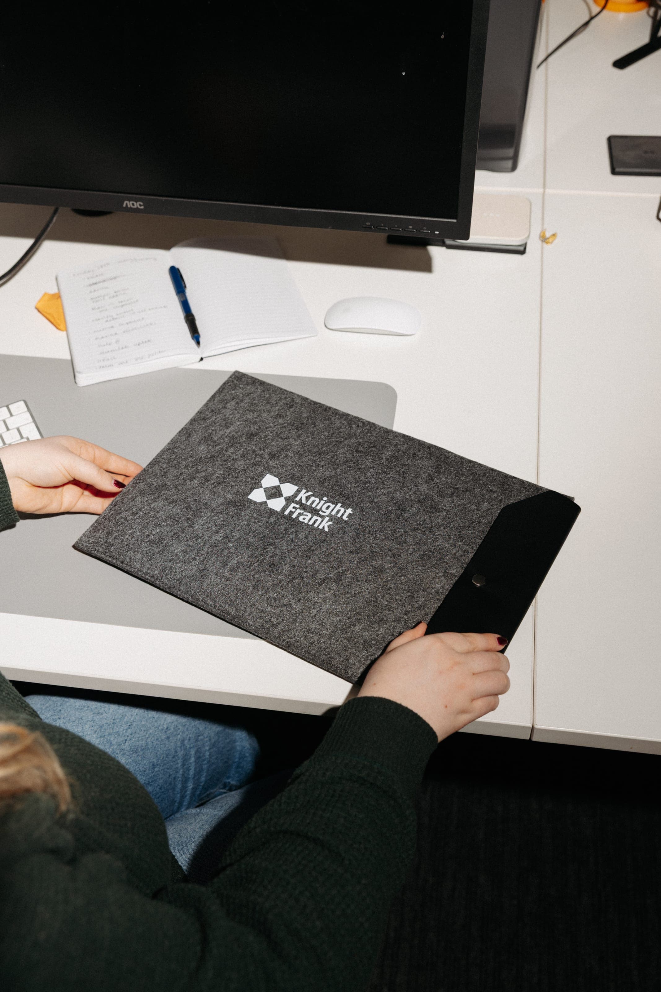 Hands hold a grey felt Knight Frank laptop sleeve with a black snap flap on a white desk