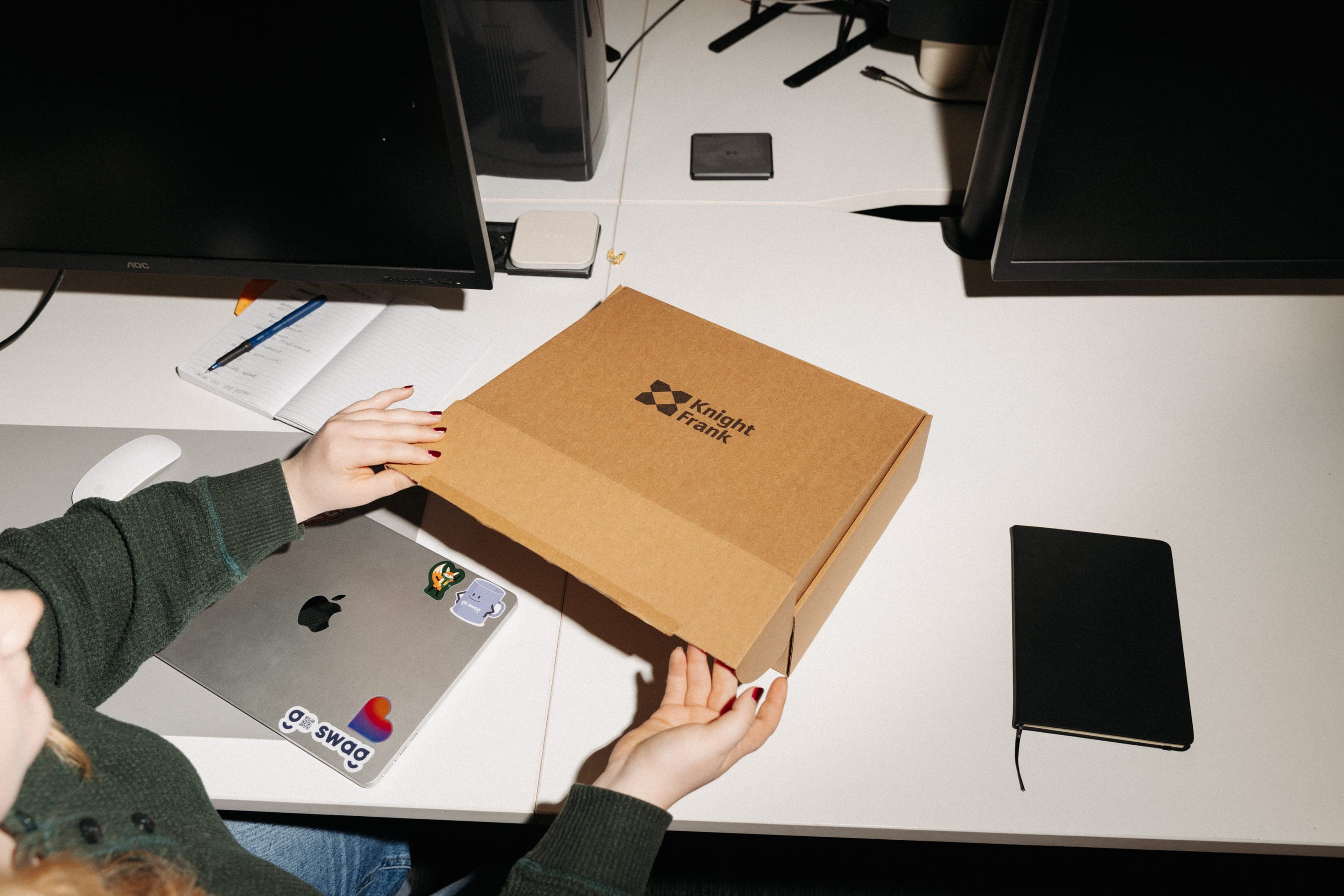 A woman at a tidy white desk lifts a kraft mailer box printed with the Knight Frank logo, beside a laptop and a black notebook