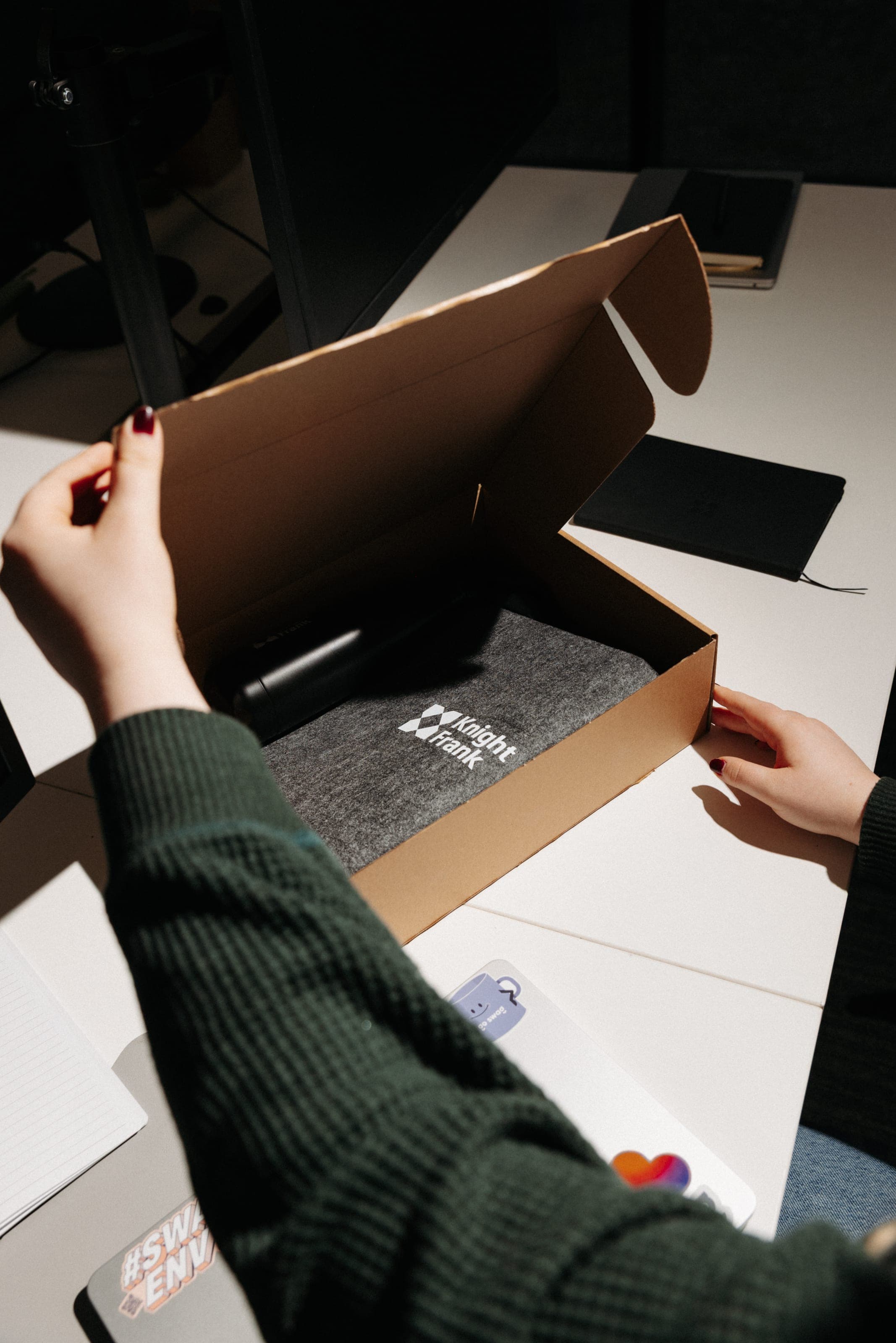 Hands open a Knight Frank kraft mailer box to reveal a grey felt Knight Frank laptop sleeve inside