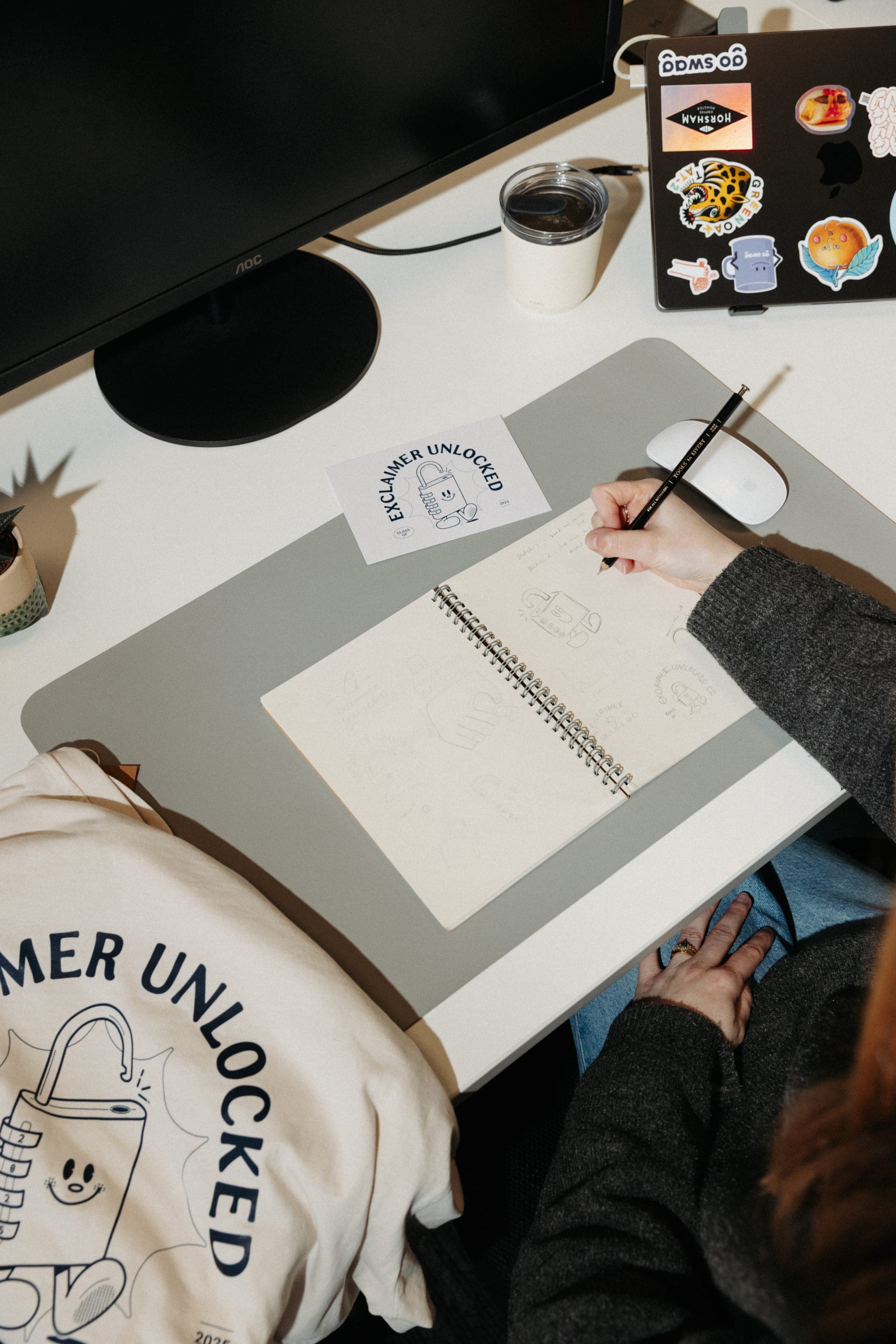 Hand sketching padlock characters in a notebook beside an 'Exclaimer Unlocked' card and cream T-shirt