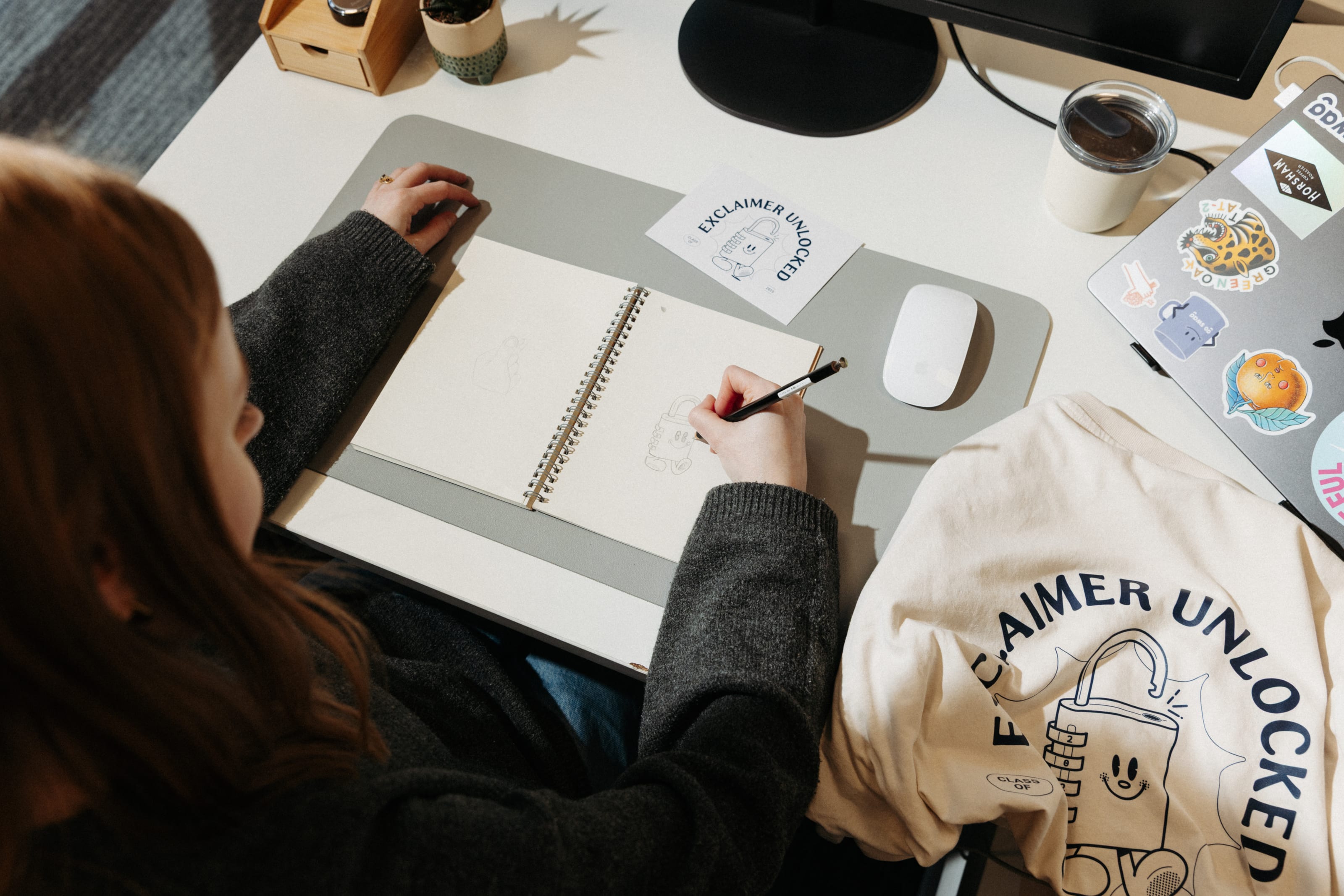 Woman sketching a padlock character at a sunlit desk beside an ‘Exclaimer Unlocked’ card and T-shirt