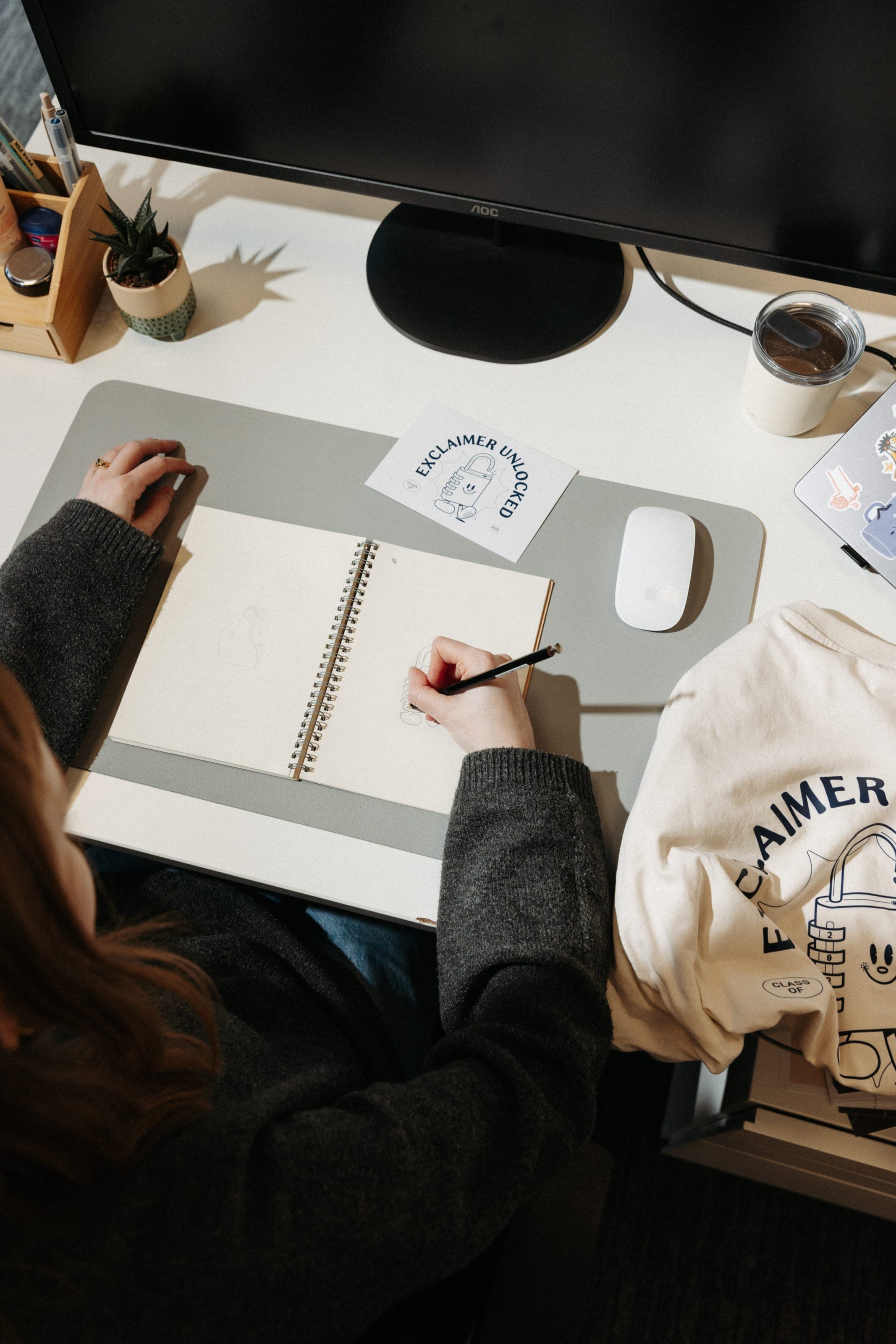 Overhead view of a designer sketching in a spiral notebook beside an Exclaimer Unlocked card and a cream Exclaimer T-shirt