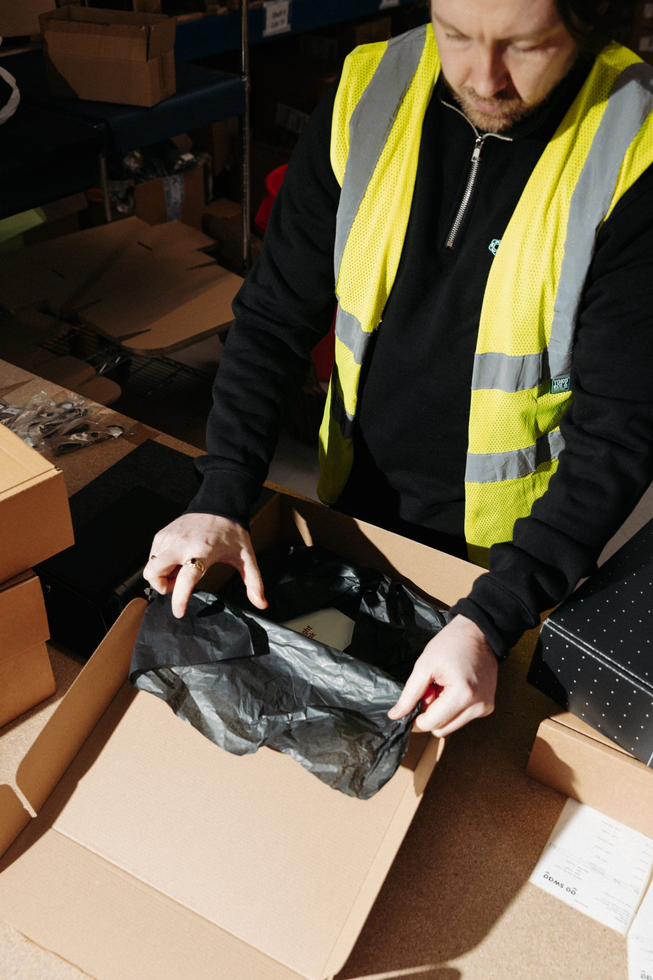 Man in a hi-vis vest folding black tissue into a kraft box at a warehouse bench