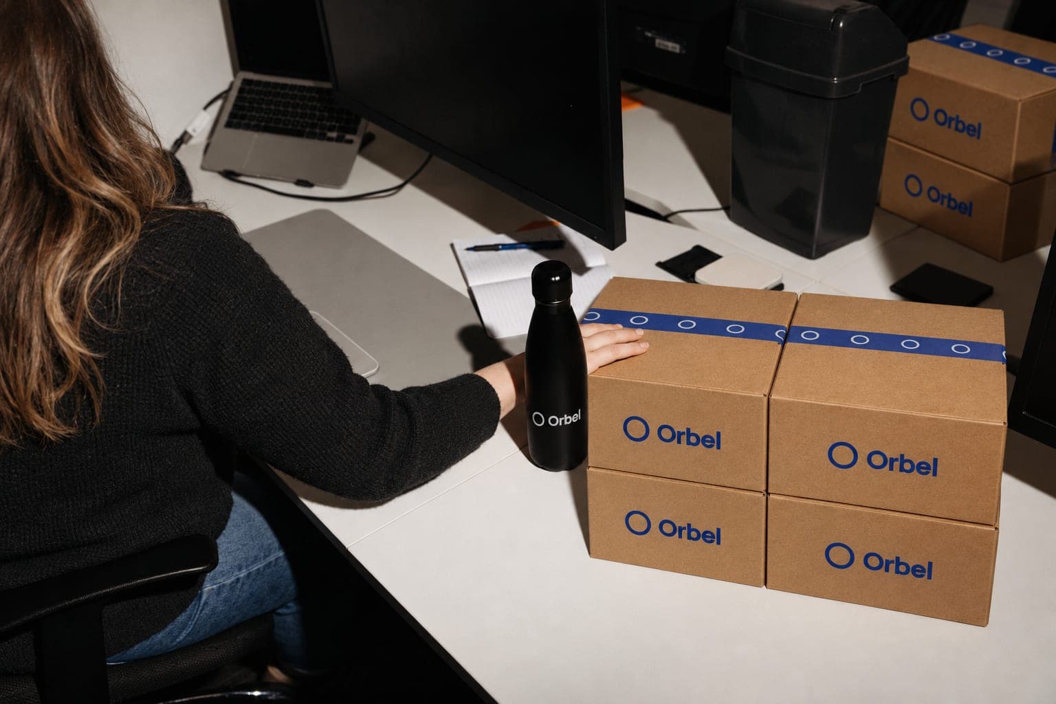 A woman at her office desk rests a hand on a neat stack of sealed, printed delivery boxes