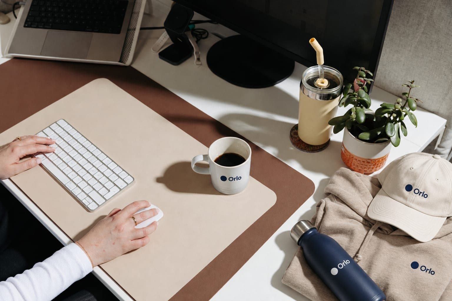 Hands at a keyboard on a tidy desk, with a branded mug of coffee, a folded hoodie, a cap and a navy bottle beside them