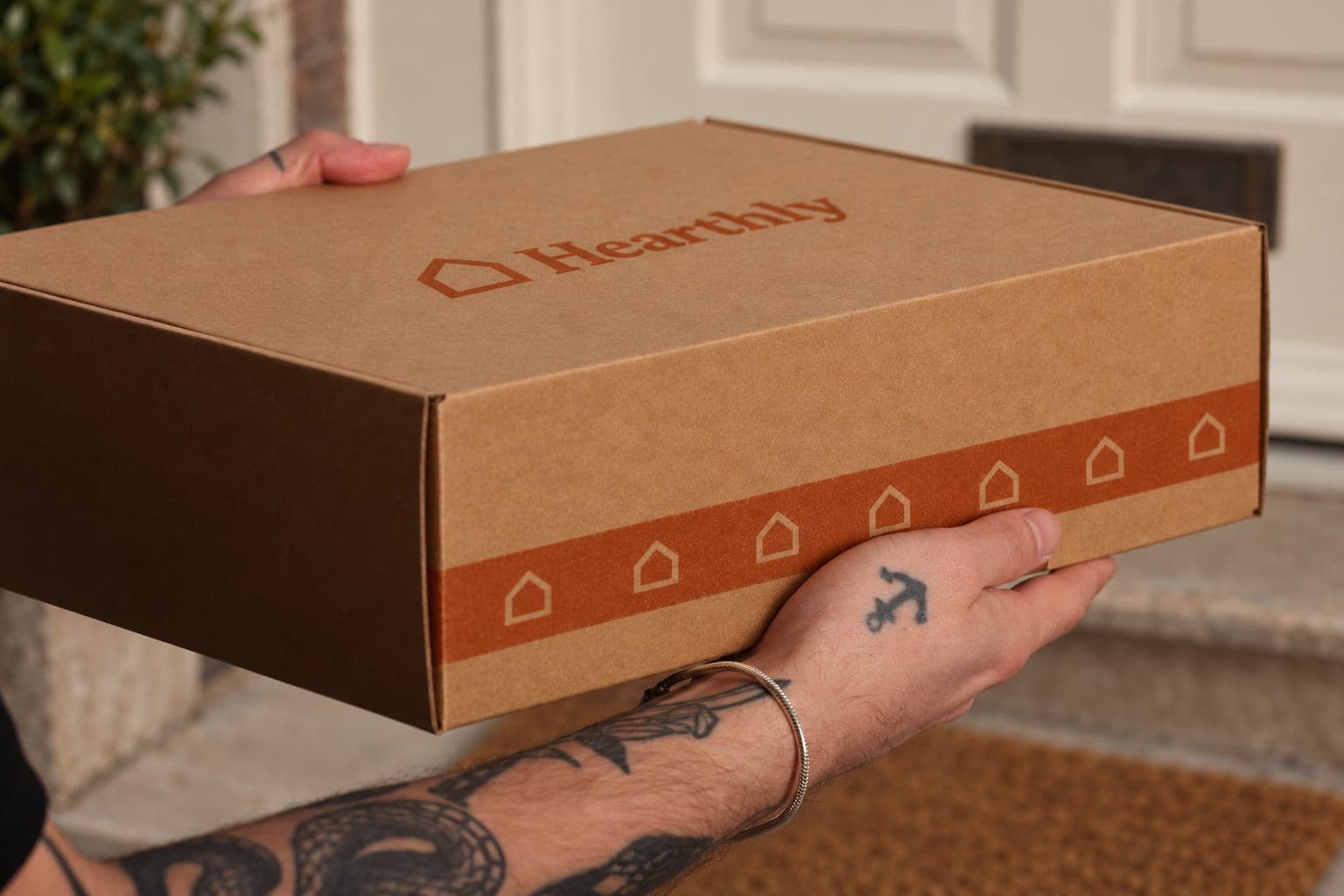 Hands hold a printed gift box at a front door, with the doormat and step behind