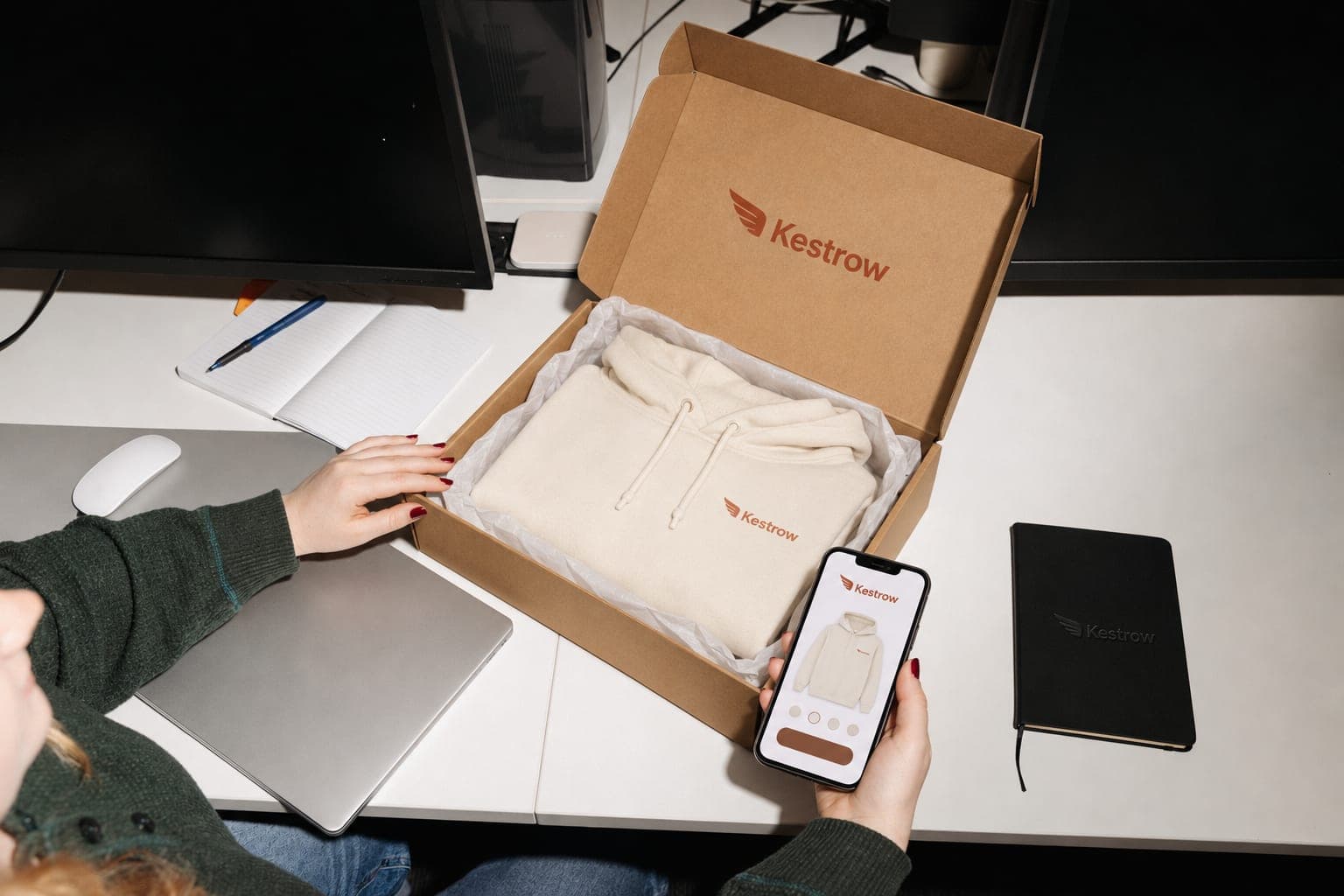 A woman holds her phone showing a gift claim page with a hoodie and size options, next to an open box with the same hoodie folded in tissue
