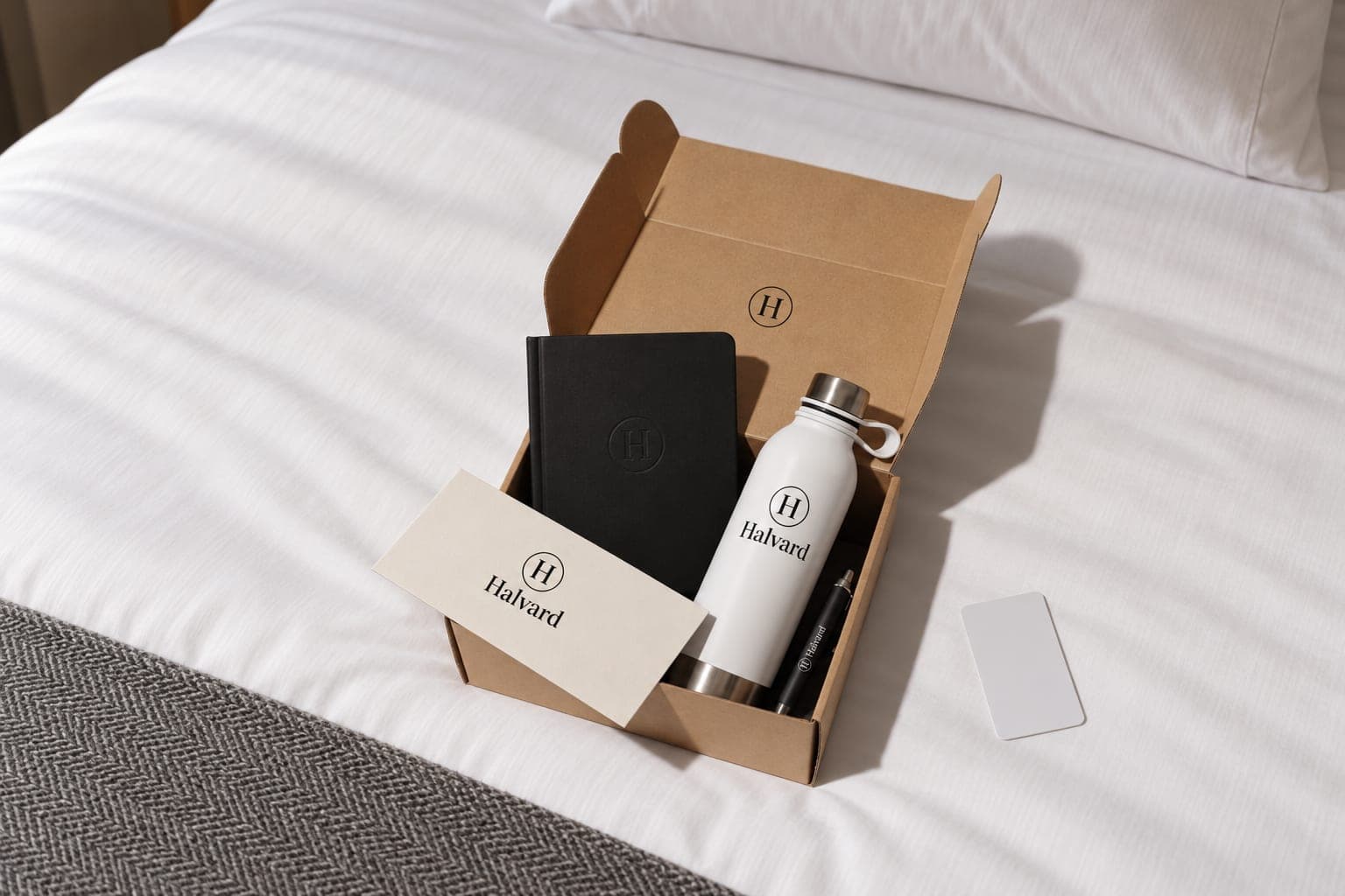 An open welcome box on a hotel bed holding a notebook, a bottle, a pen and a welcome card, with a key card beside it