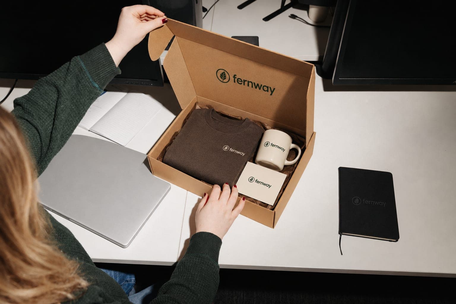 A woman at her desk opens a gift box holding a folded sweatshirt, a mug and a card, with a matching notebook beside it