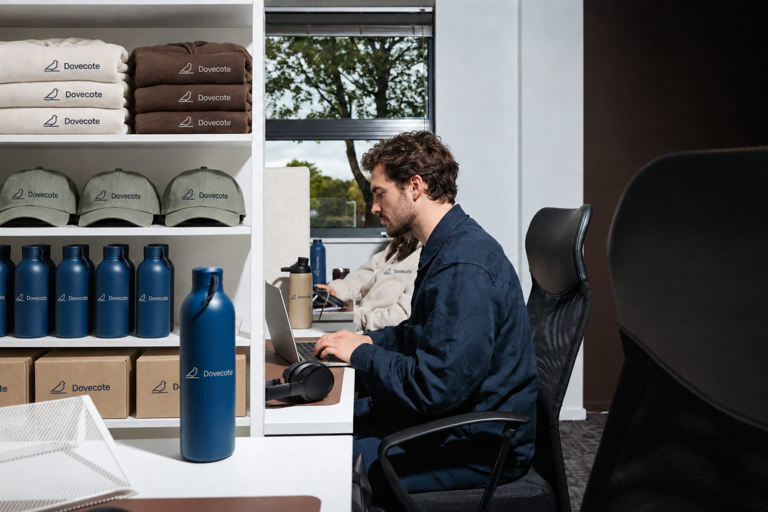 A man works at his laptop beside office shelves stocked with branded hoodies, caps, bottles and boxes