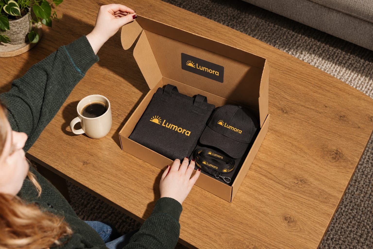 A woman at home opens a box on her coffee table holding a tote, a cap and a lanyard, with a mug of coffee beside it