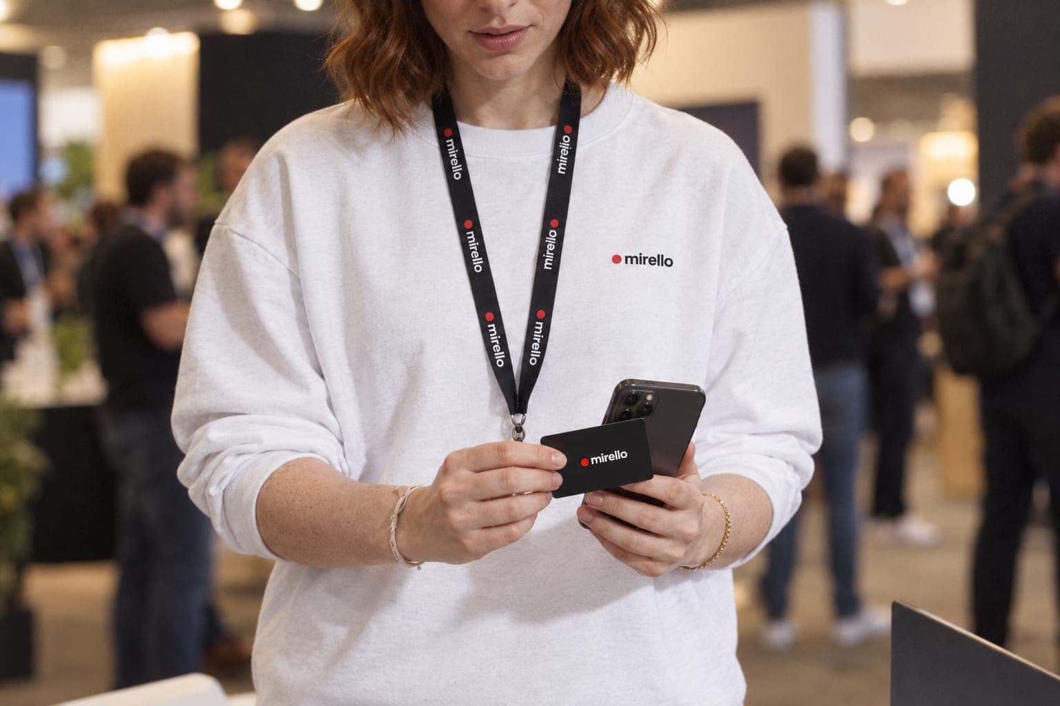 A woman at a conference taps a card against the back of her phone, with the busy exhibition hall blurred behind her