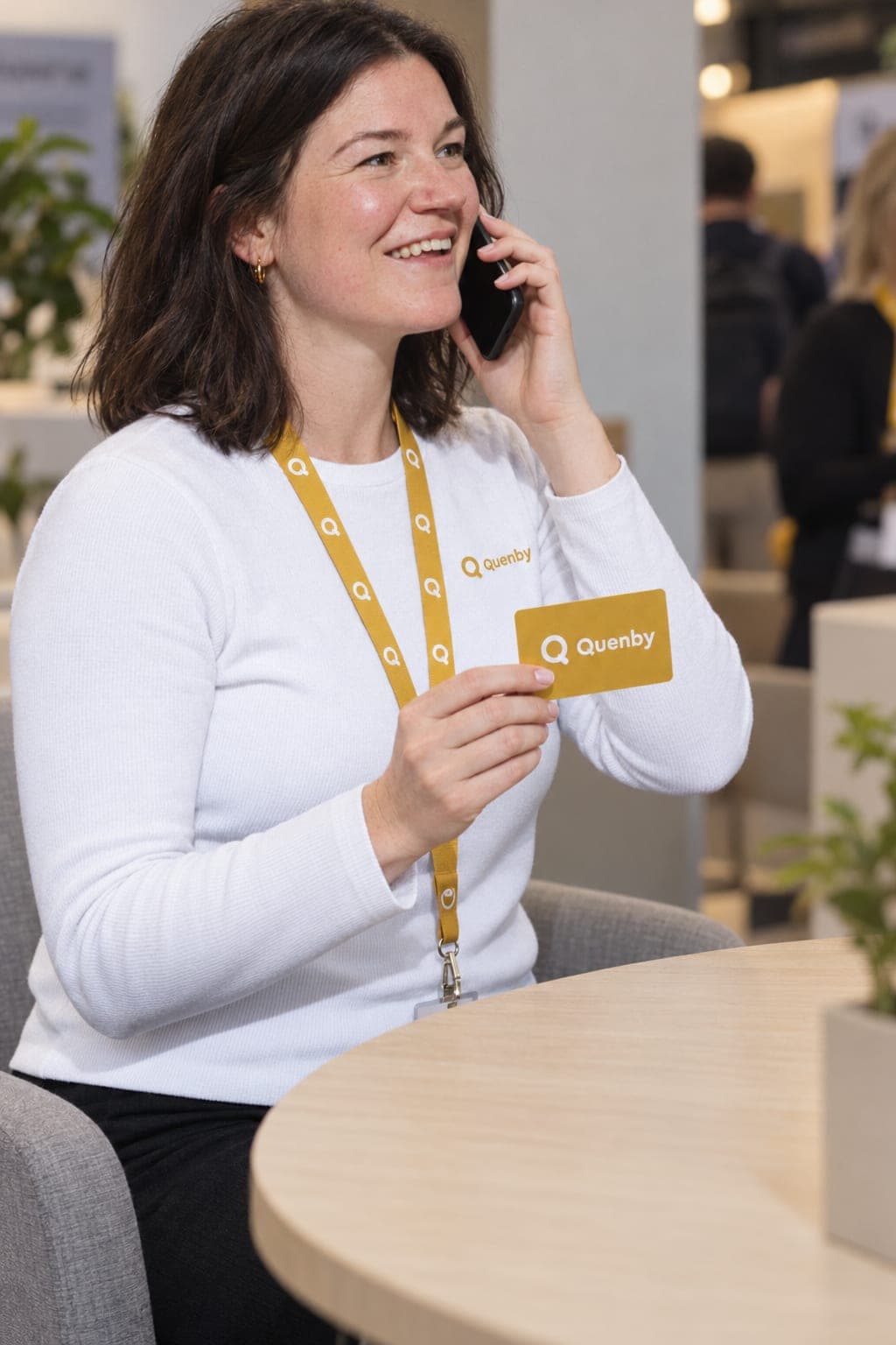 A smiling woman on a phone call at a conference lounge table holds up a yellow gift card