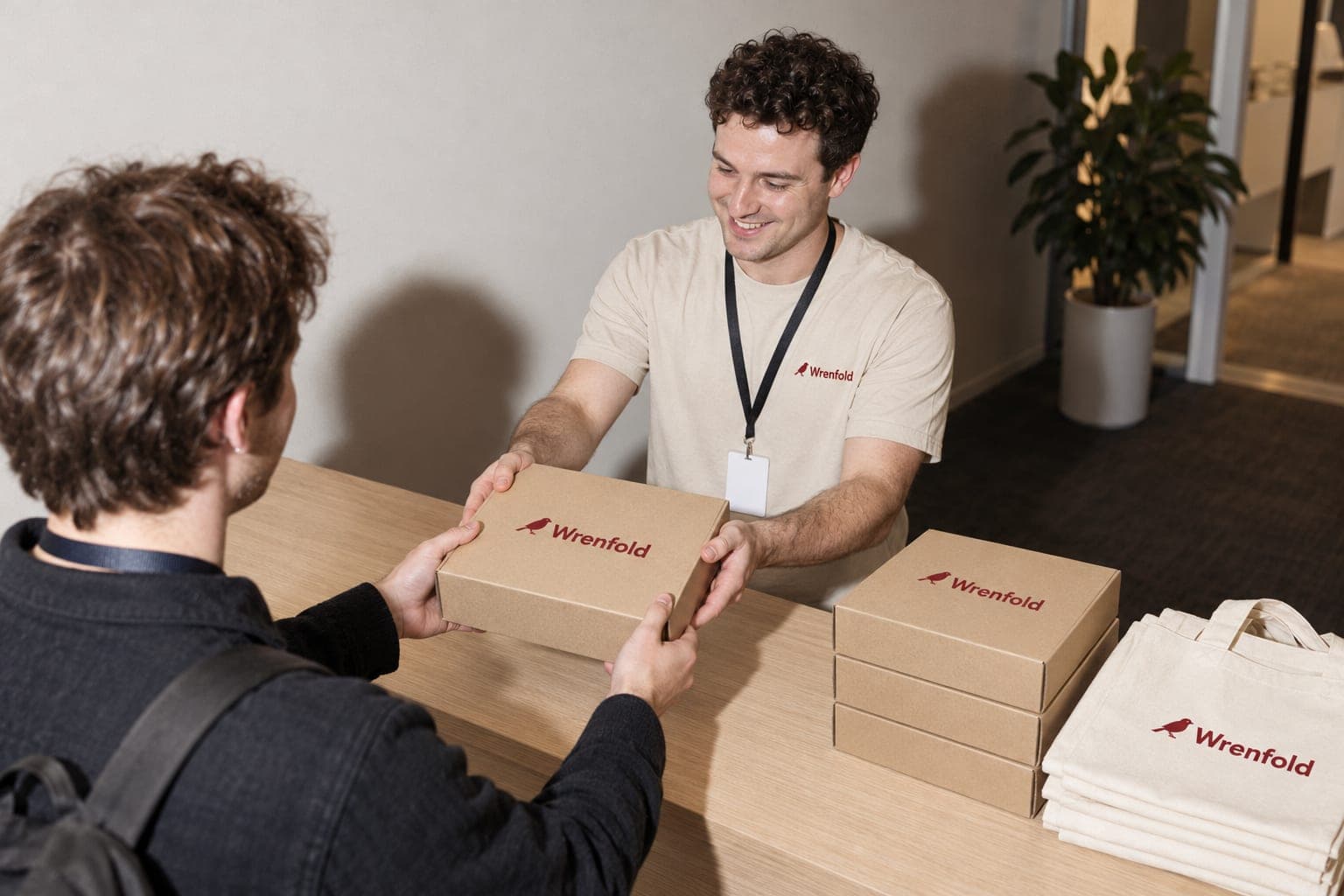A smiling team member at a welcome counter hands a box to an arriving guest, with more boxes and folded tote bags beside them