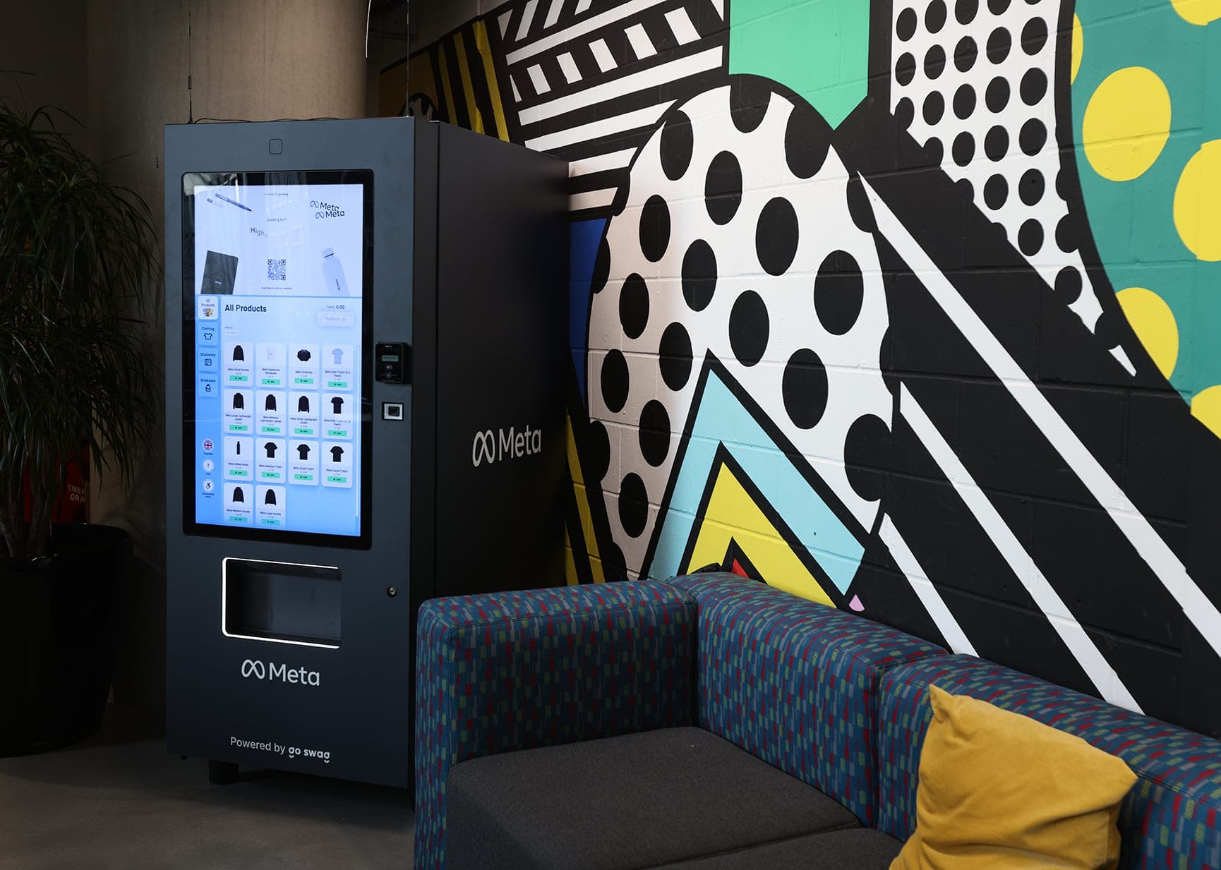 A dark Meta vending machine with a touchscreen of swag, marked “Powered by go swag”, beside a bold mural and a patterned sofa