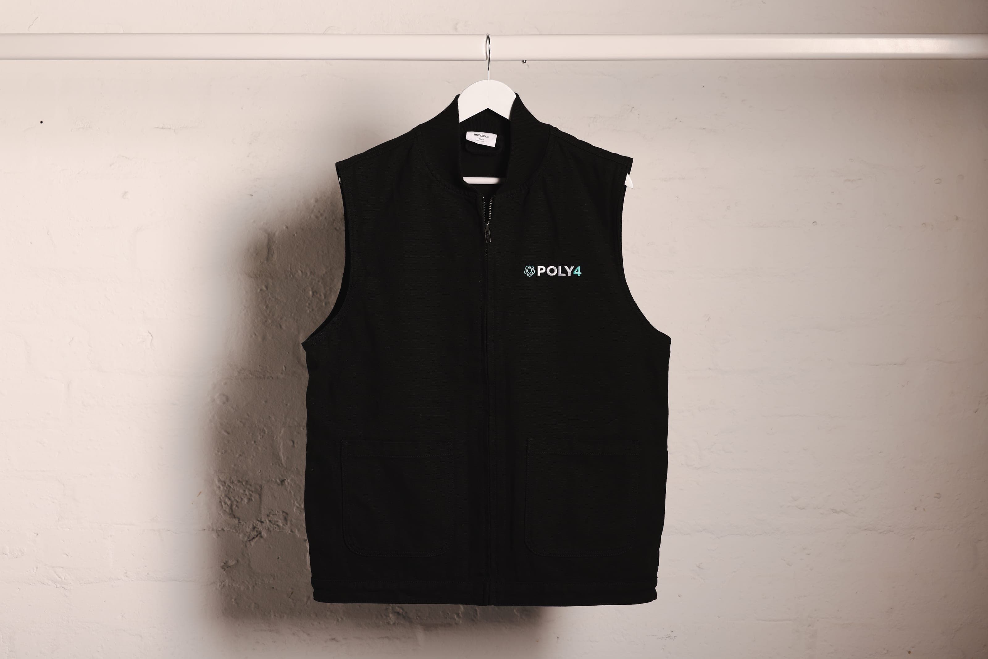 A black zip gilet with POLY4 chest embroidery on a white rail against a whitewashed brick wall