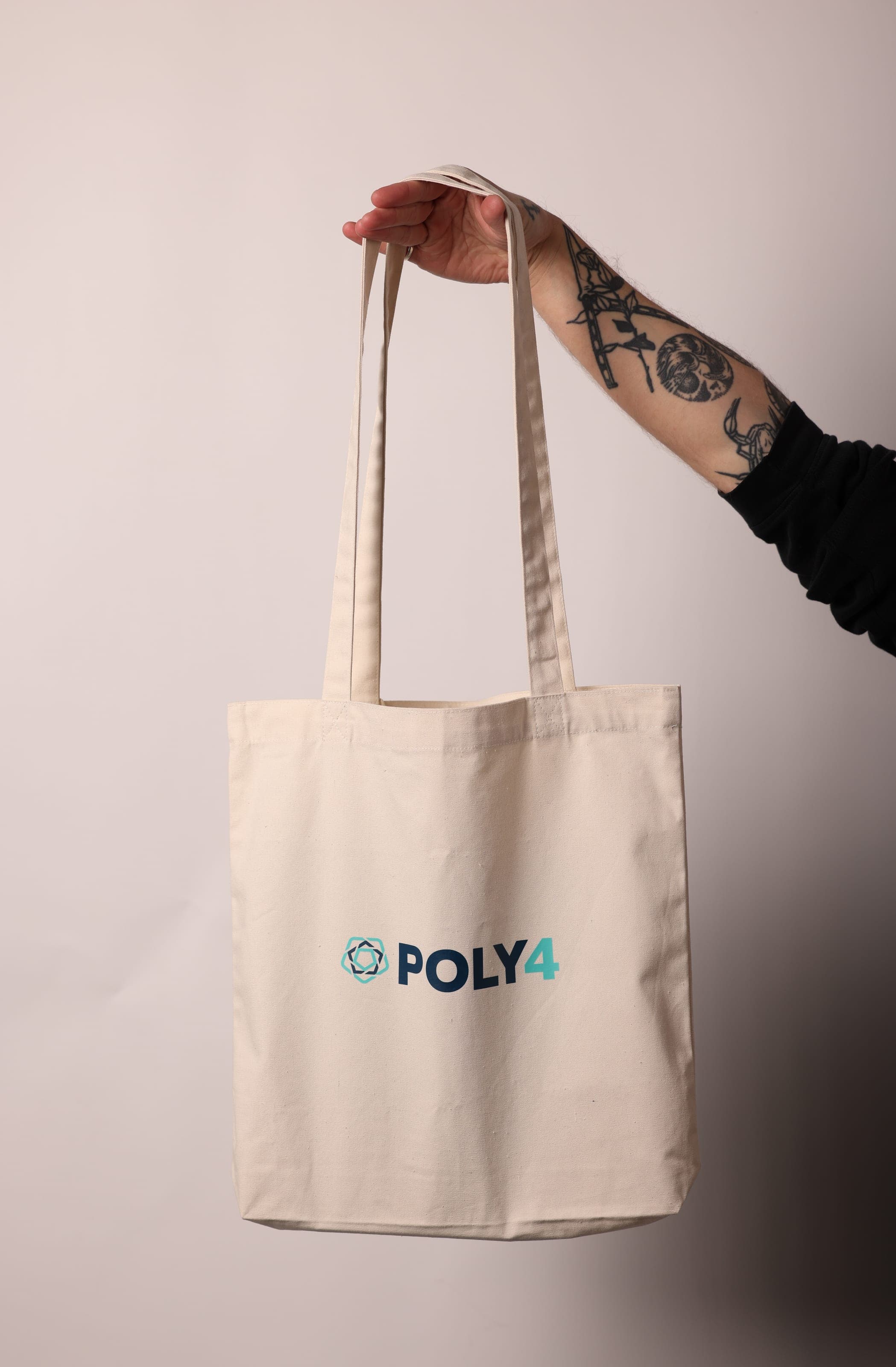 A tattooed arm holds out a natural canvas tote printed with the POLY4 logo