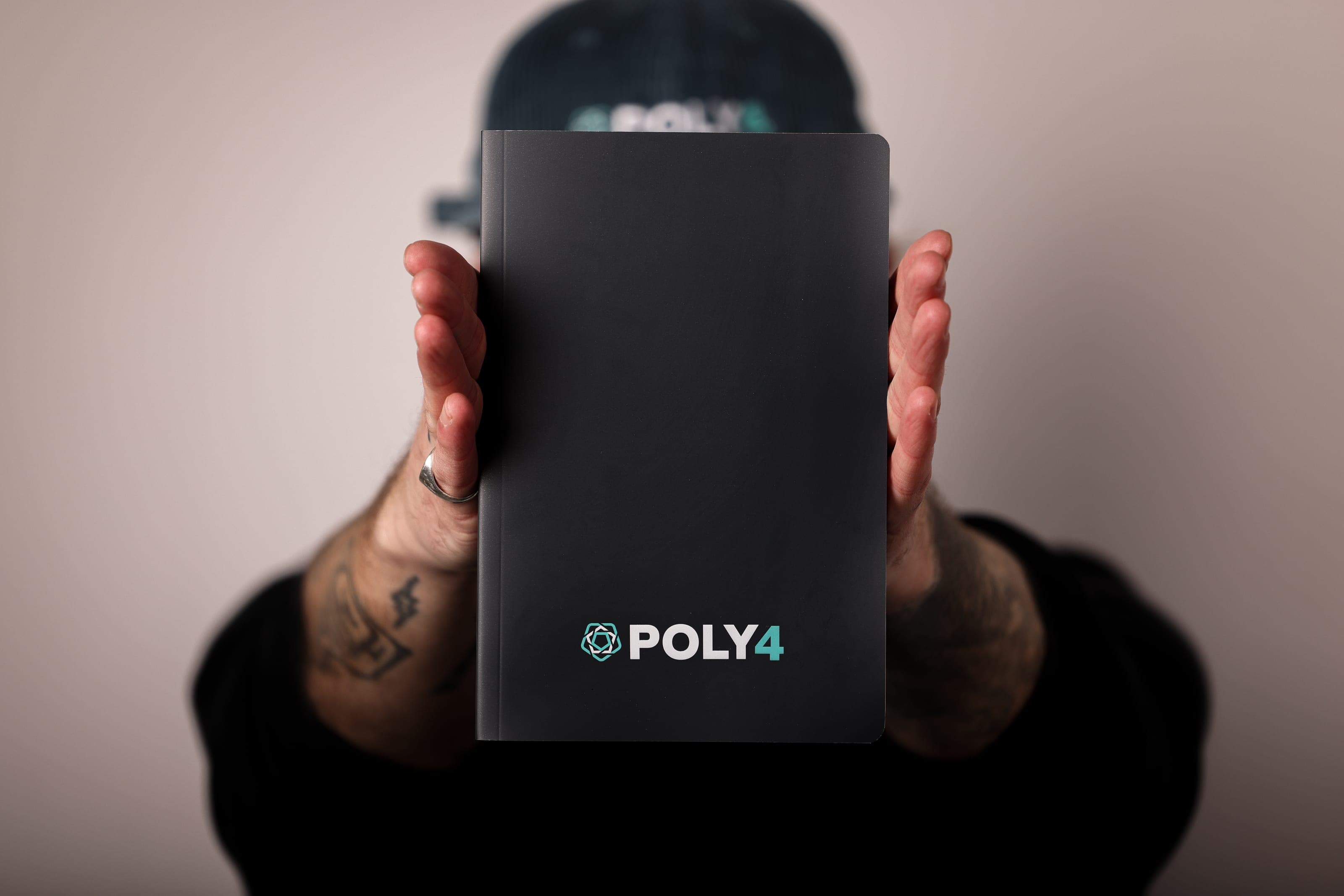 Tattooed hands hold a black POLY4 notebook up in front of a face