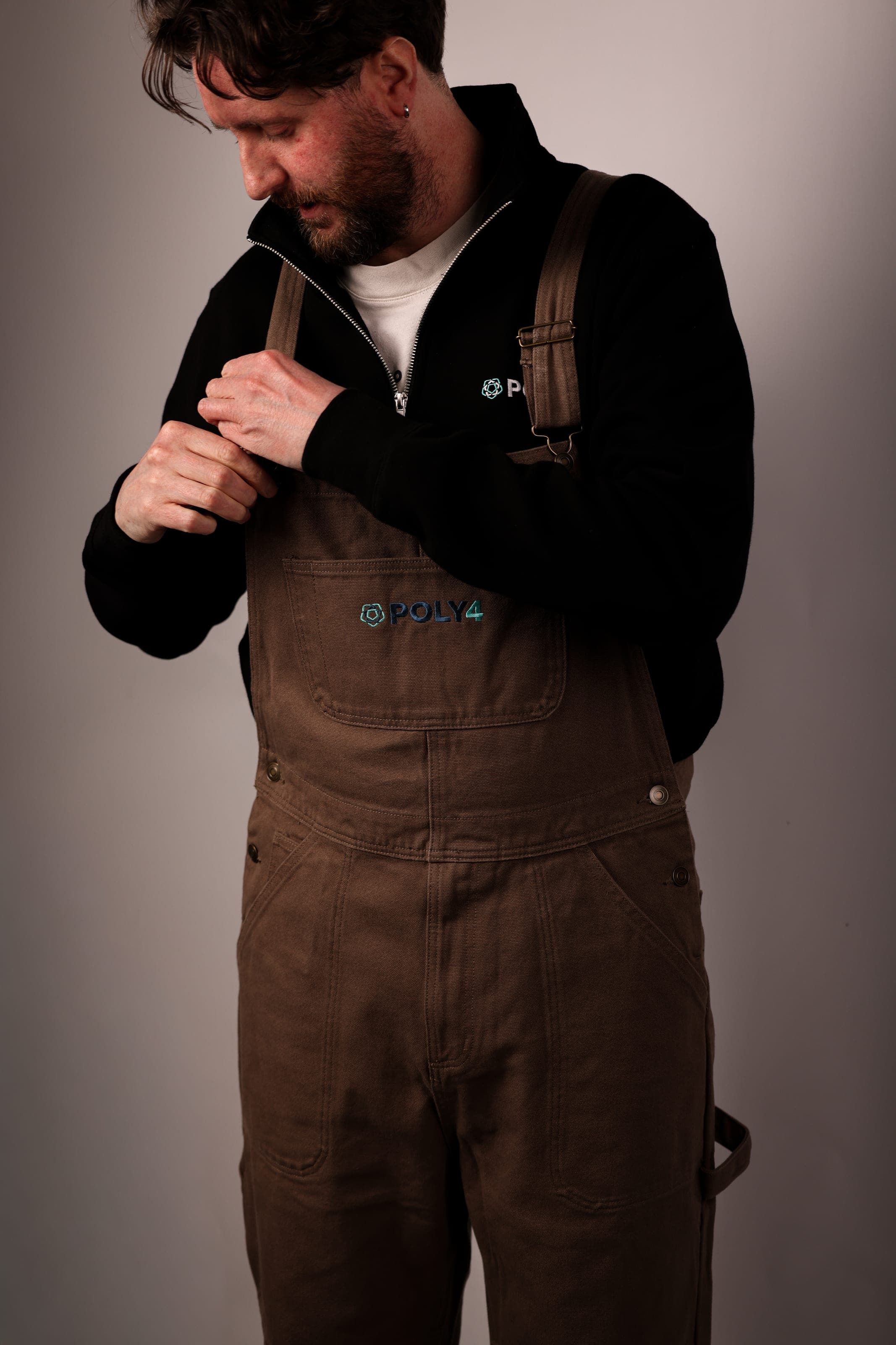 A man adjusting brown POLY4 work overalls worn over a black POLY4 quarter-zip