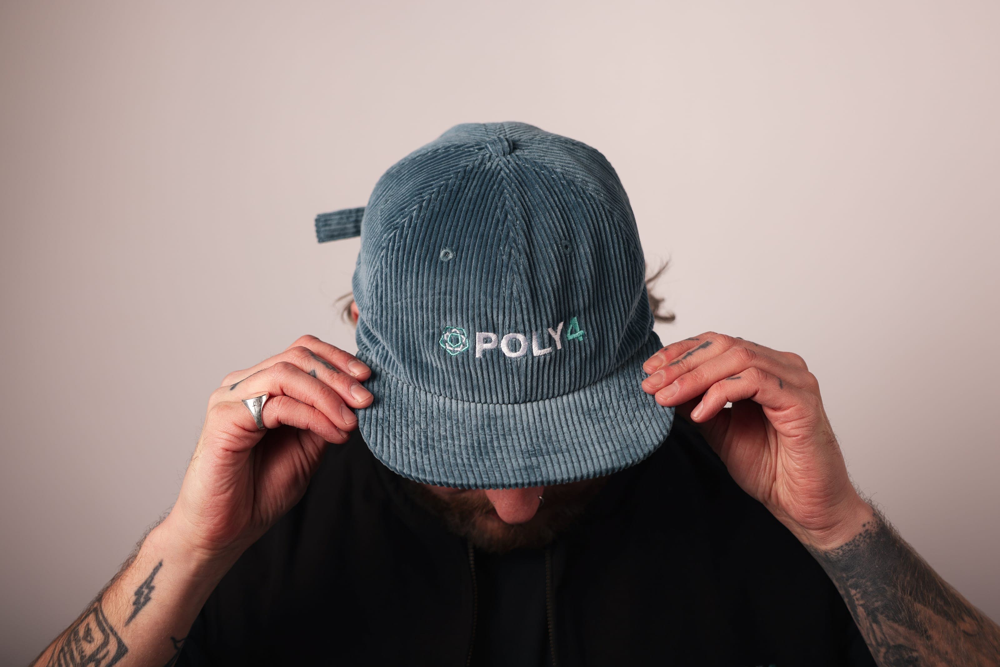 A man pulls a blue-grey corduroy cap embroidered with POLY4 down over his face