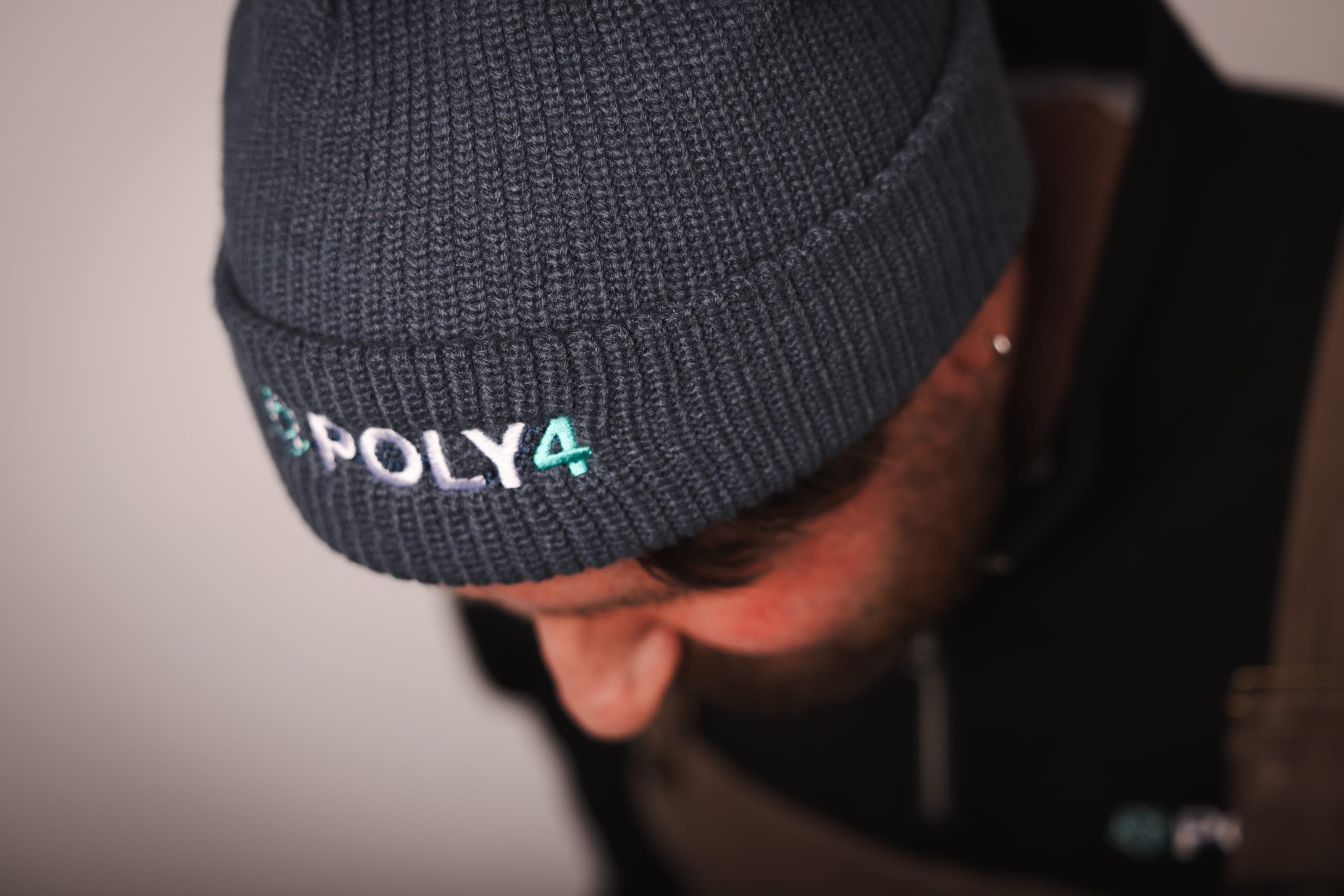 Close-up of a charcoal ribbed beanie embroidered with POLY4 in white and teal, worn by a man looking down