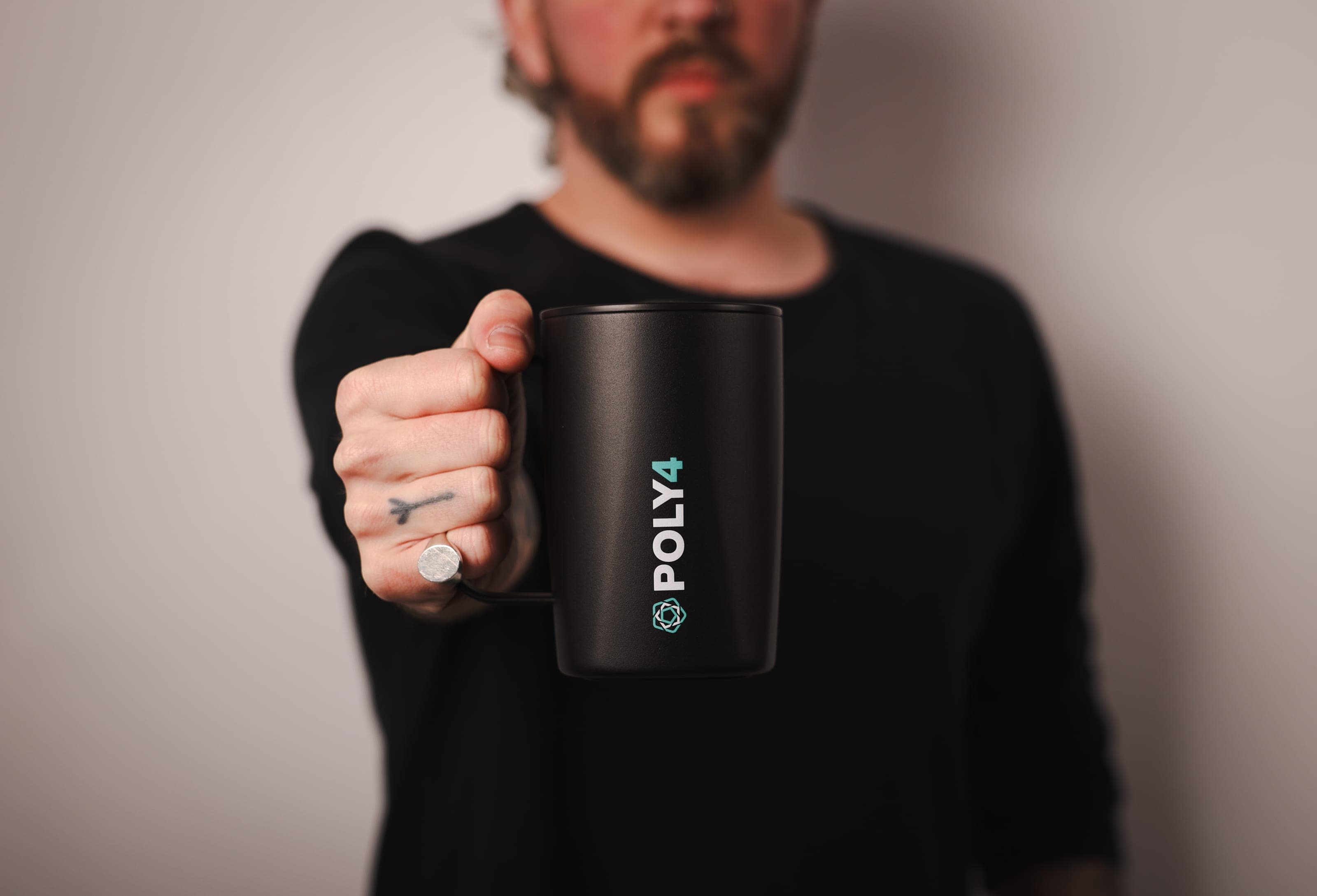 A bearded man in black holds a matte black POLY4 mug towards the camera