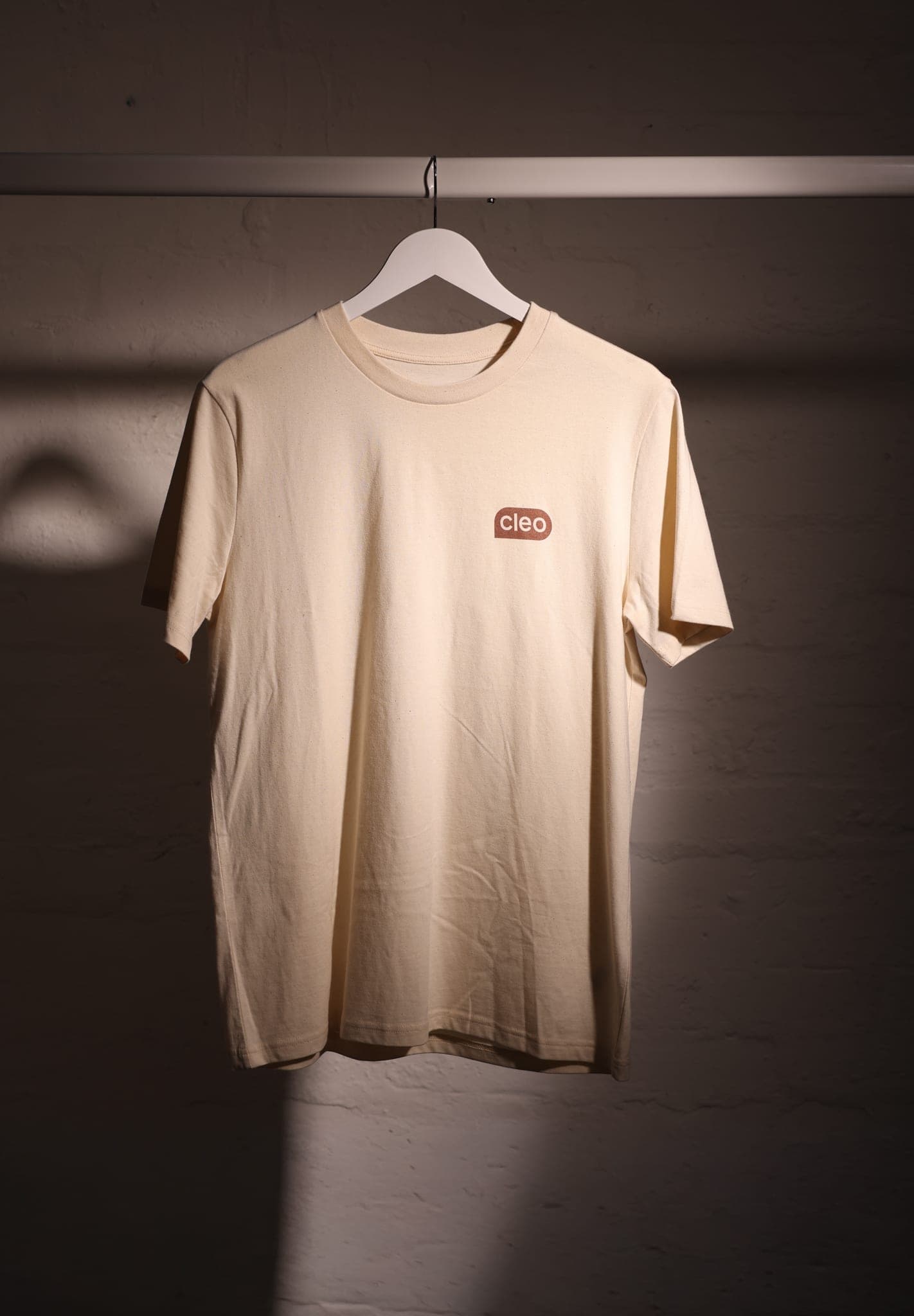 Cream T-shirt with a small brown Cleo logo on a hanger in a shaft of light