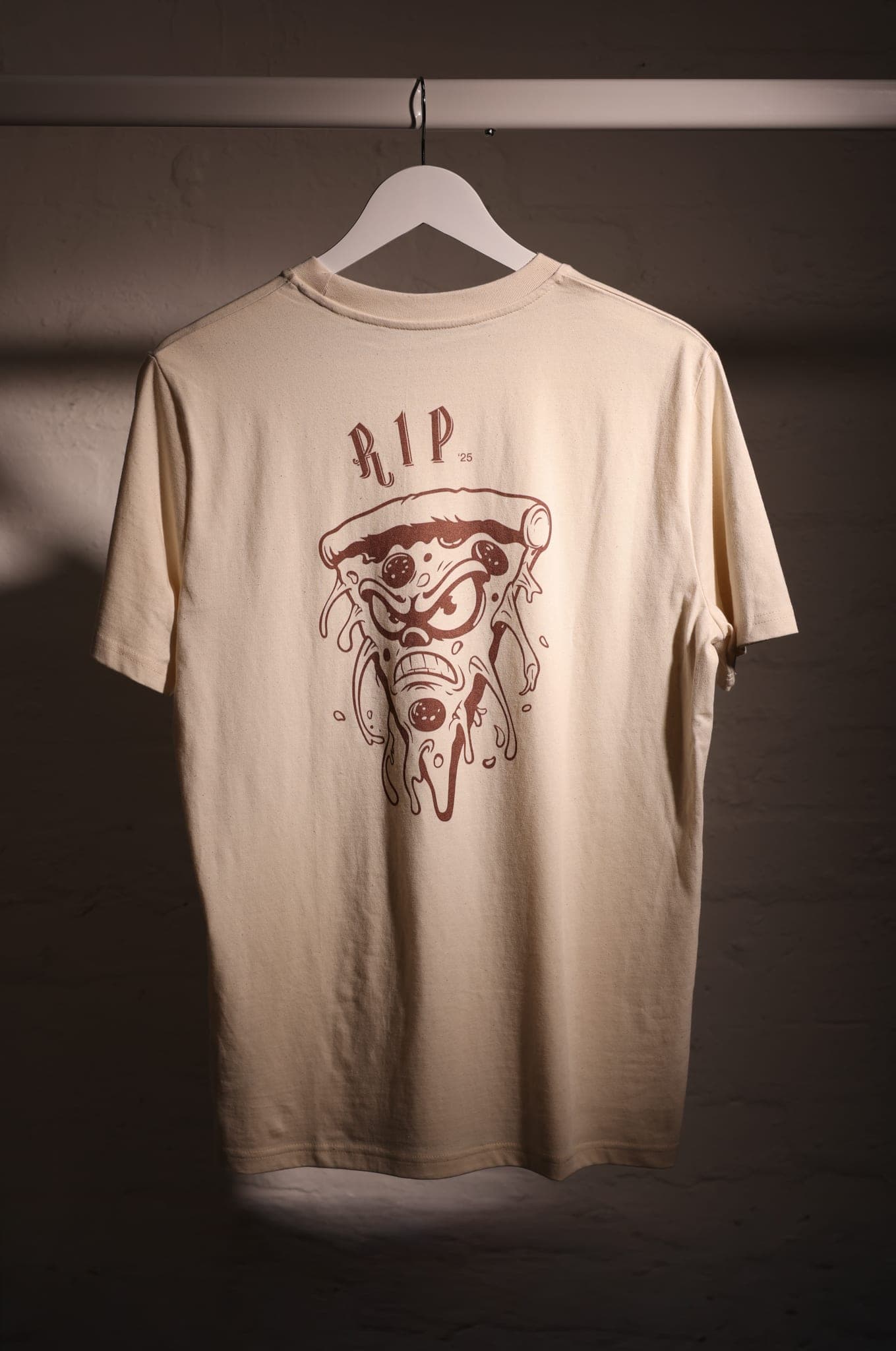 Back of a cream T-shirt with a brown illustrated pizza slice and the words RIP ’25