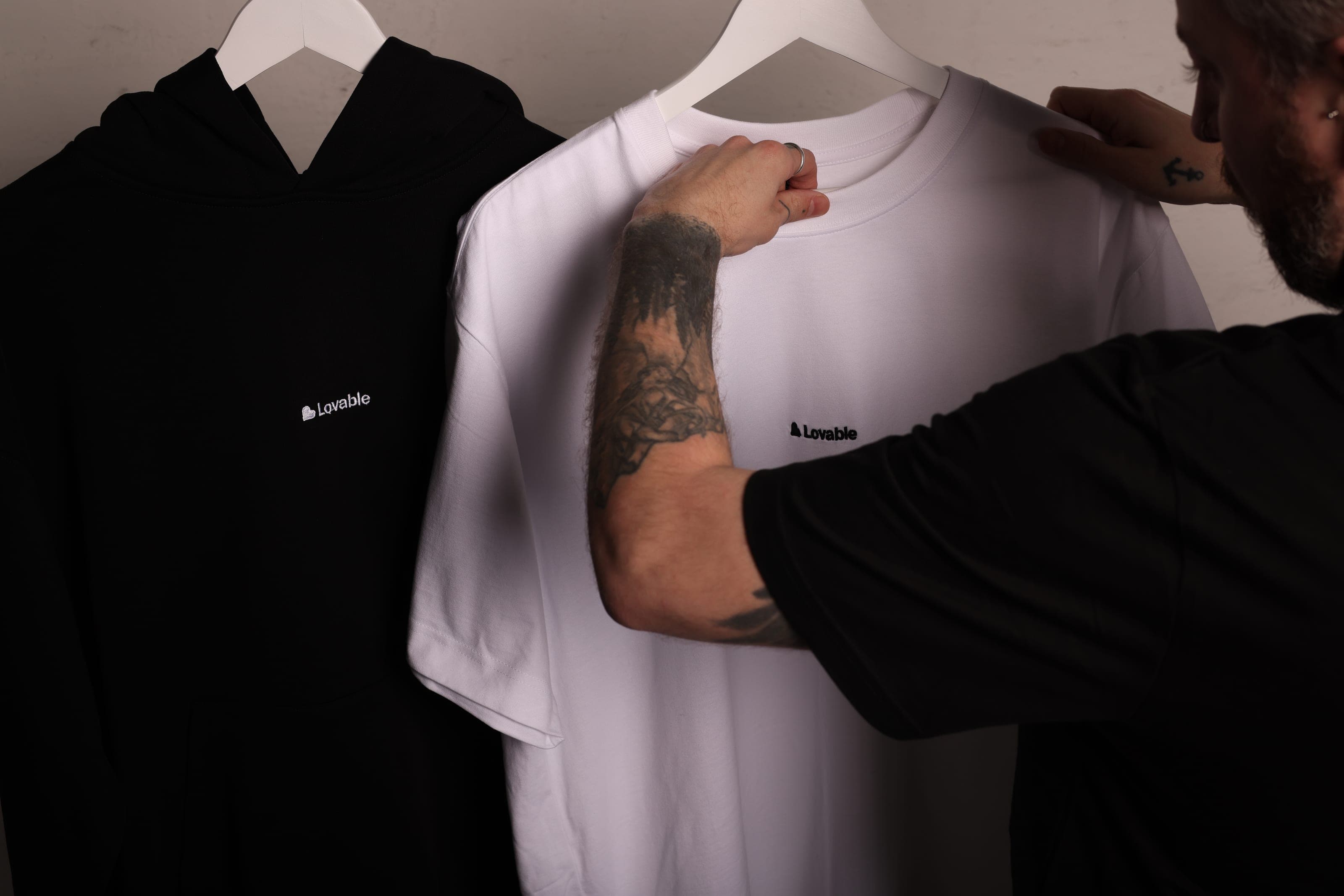 A man holds up a white Lovable T-shirt beside a black Lovable hoodie
