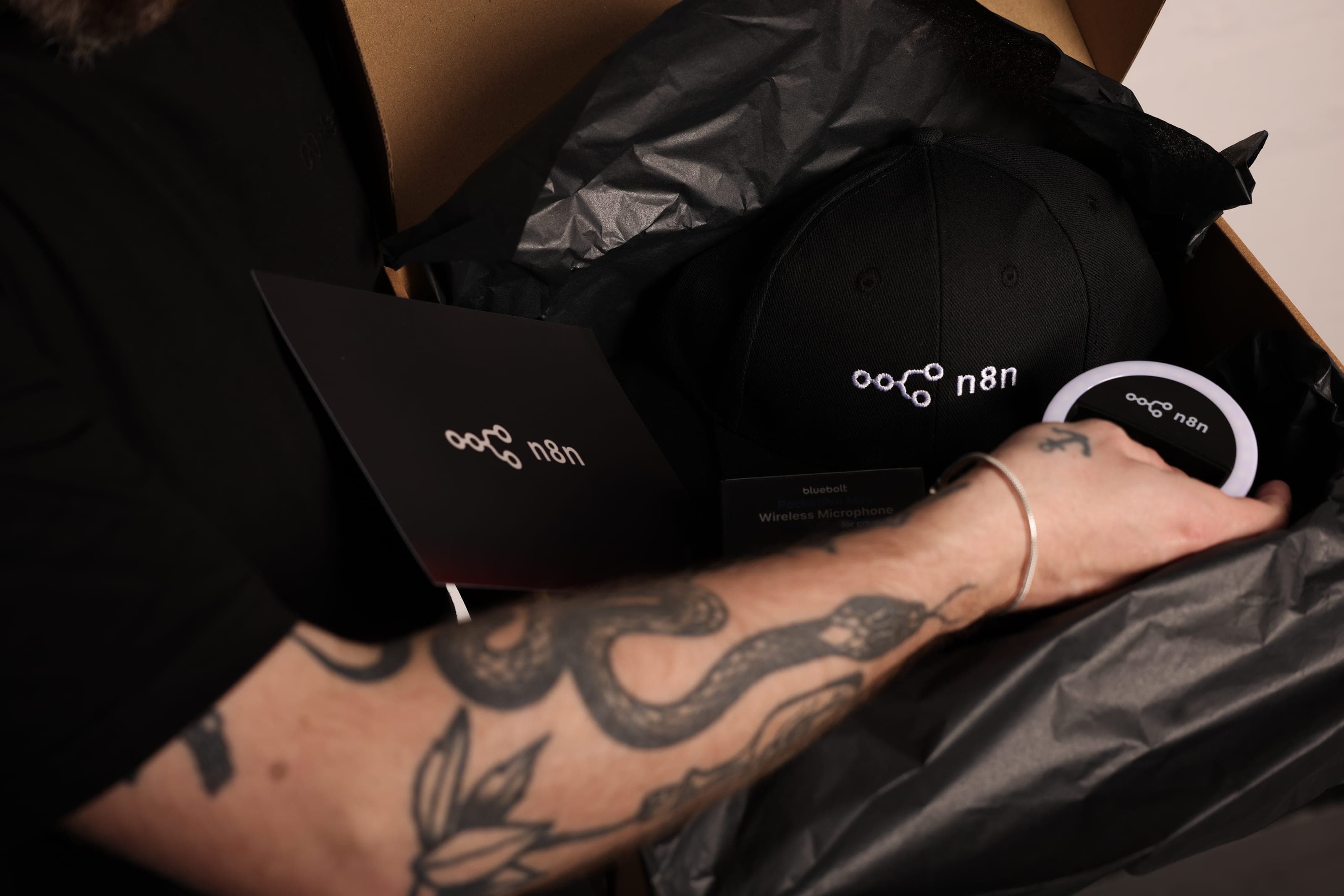 An arm reaches into an opened box of black tissue holding an n8n cap, an n8n card and a round n8n accessory