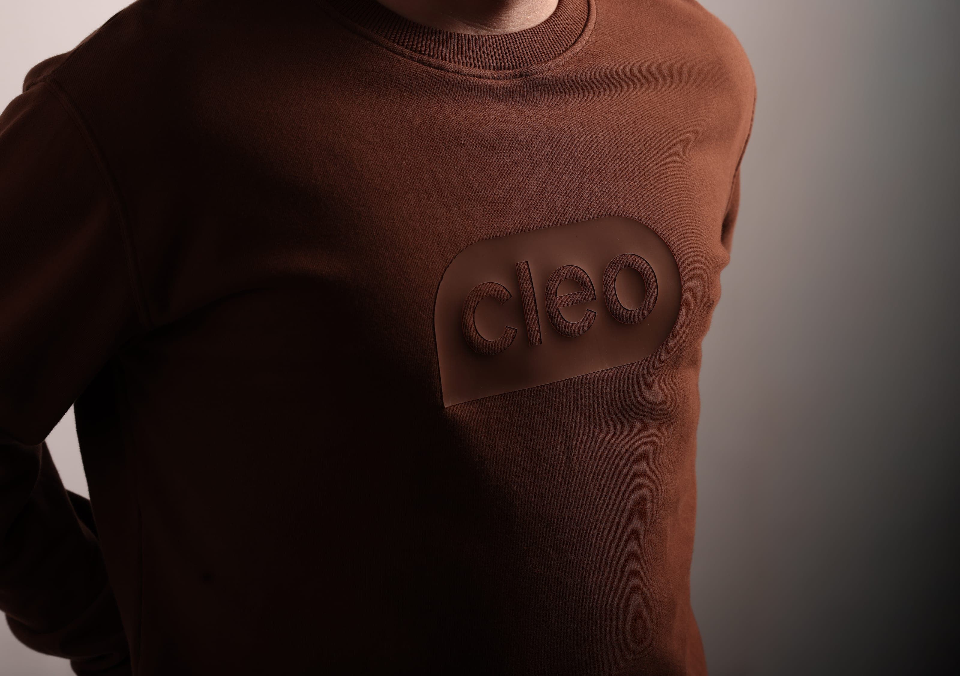 Brown sweatshirt with a tonal raised Cleo logo patch on the chest, lit from the side