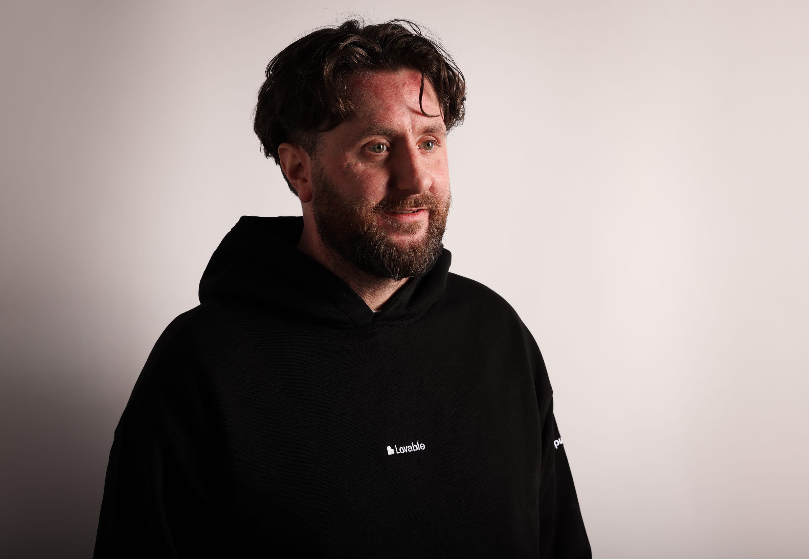 A bearded man with dark hair in a black Lovable hoodie looks off to the side