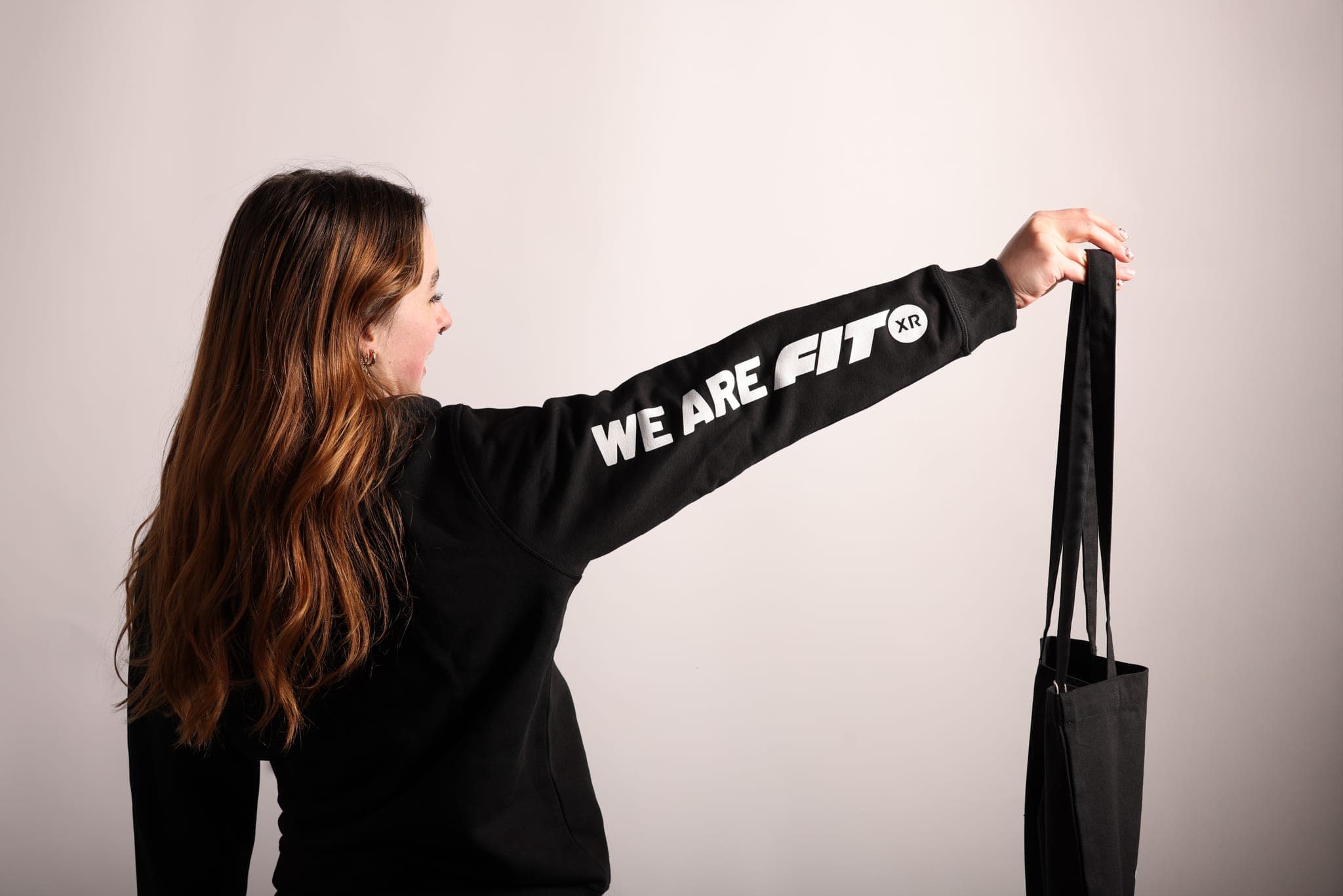 A woman holds a black tote at arm’s length, showing WE ARE FIT XR printed down her sleeve