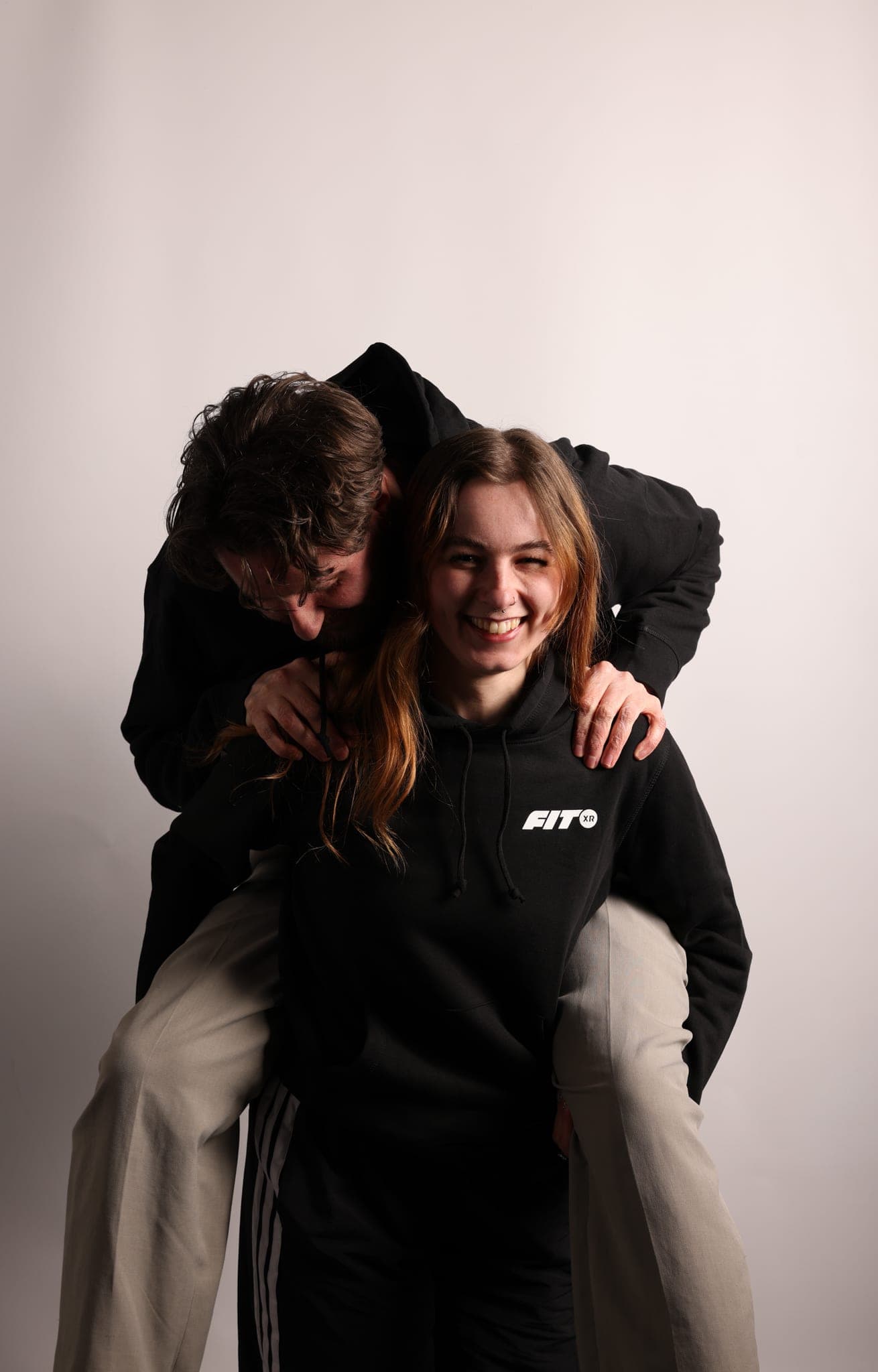 A laughing woman in a black FitXR hoodie gives a piggyback to a dark-haired man