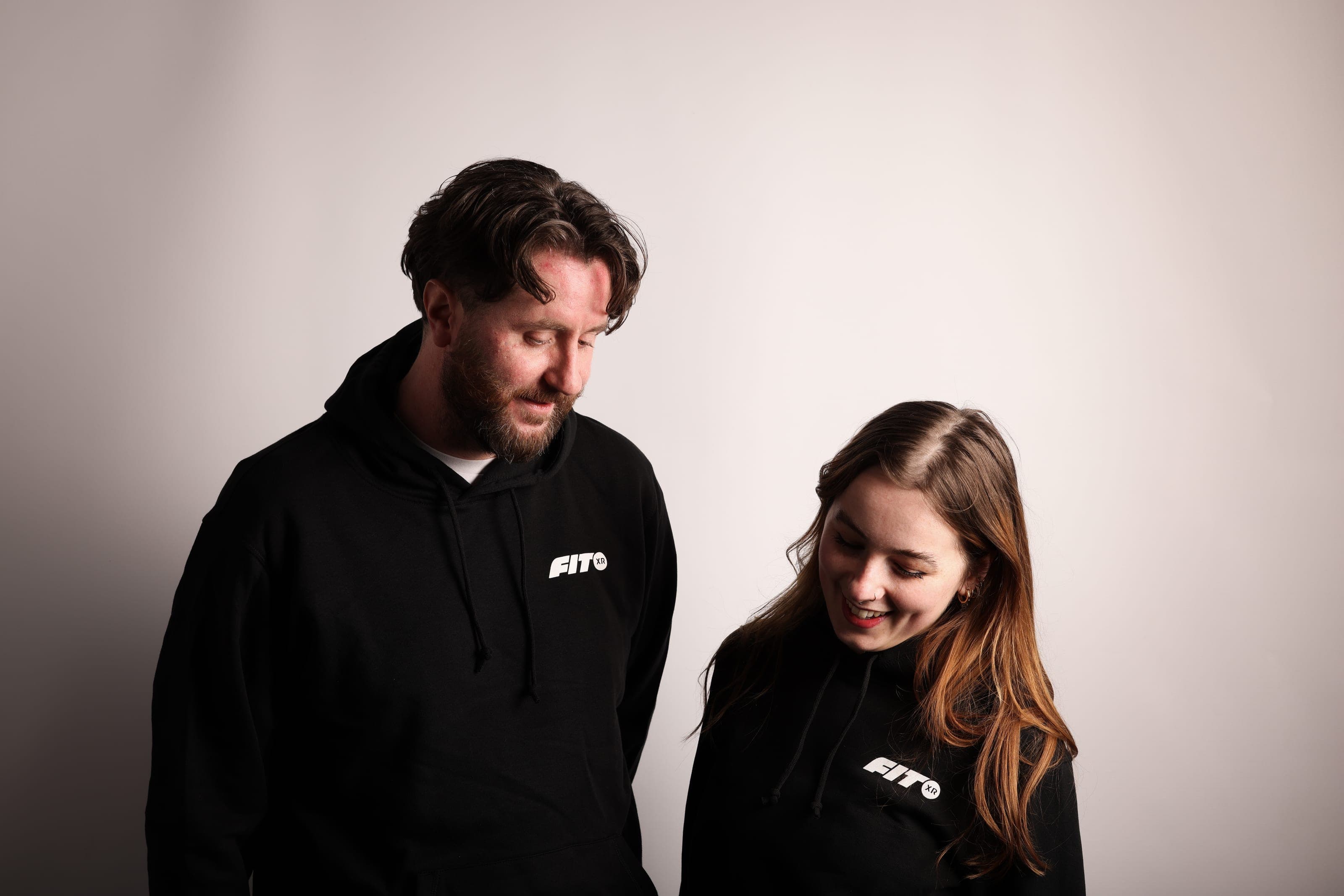 A bearded man and a woman with auburn hair, both in black FitXR hoodies with the logo on the chest, look down and smile