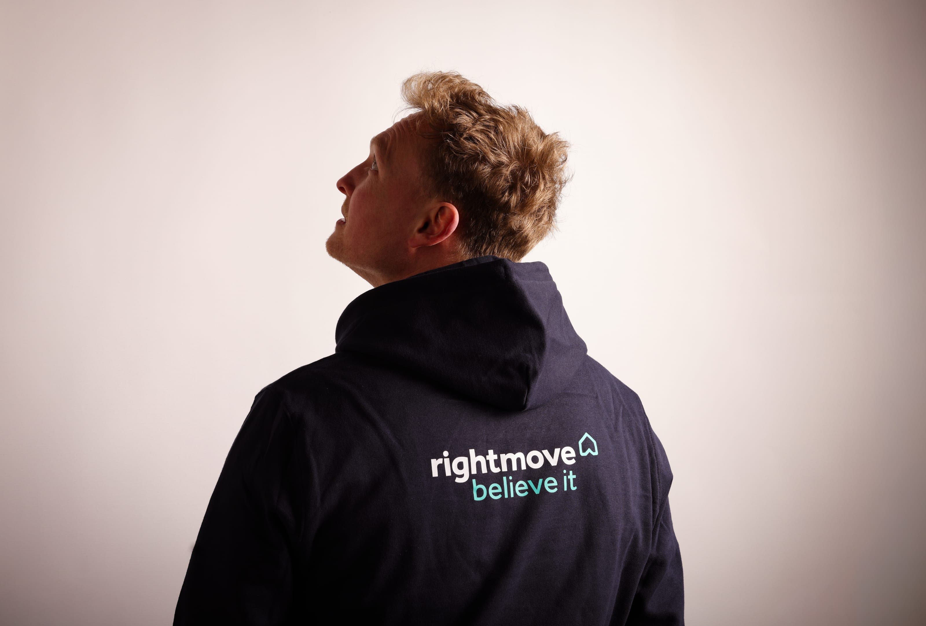 Back of a navy hoodie printed with the Rightmove logo and “believe it”, worn by a man looking up