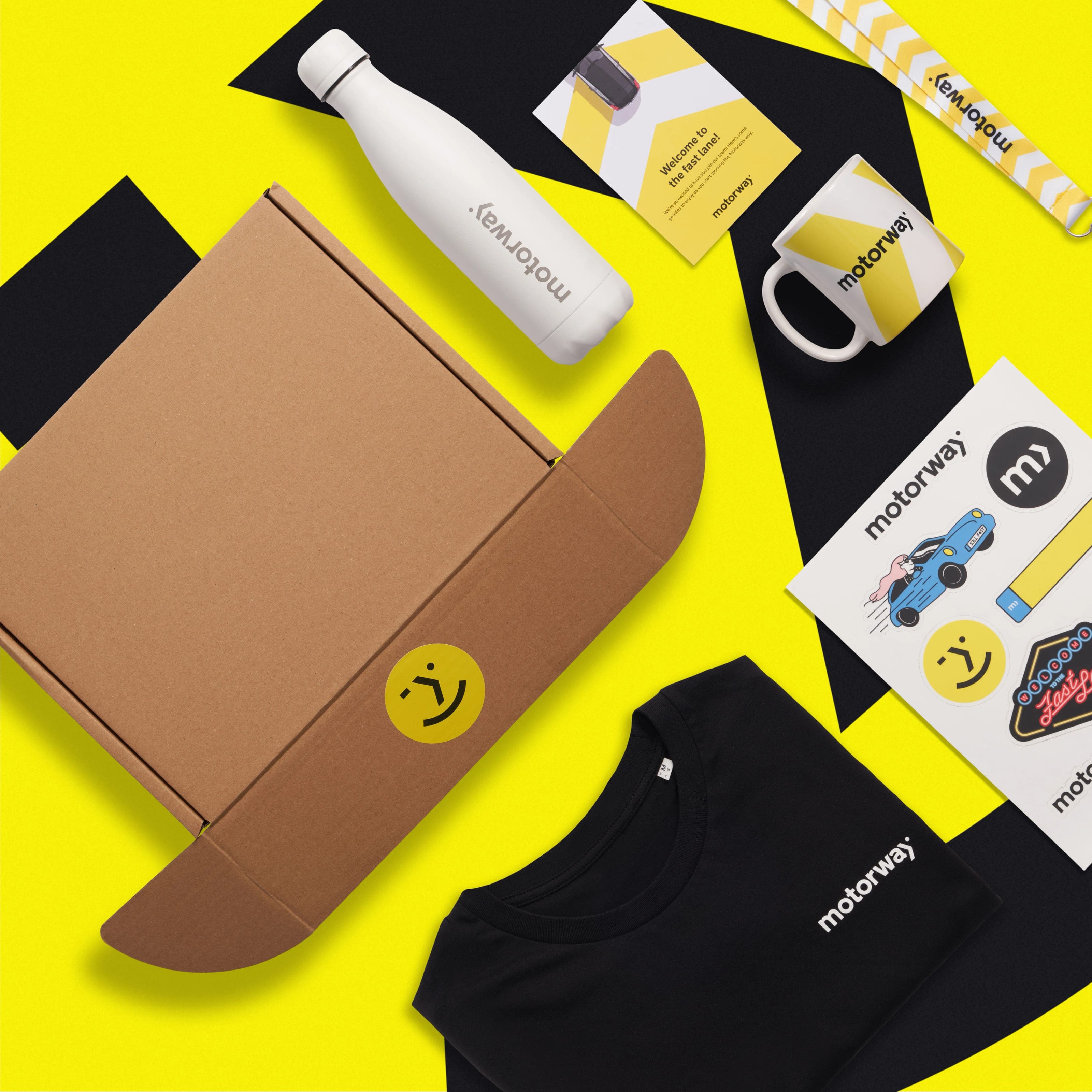Angled view of the Motorway welcome kit: kraft box, white bottle, welcome card, chevron mug, stickers and black T-shirt