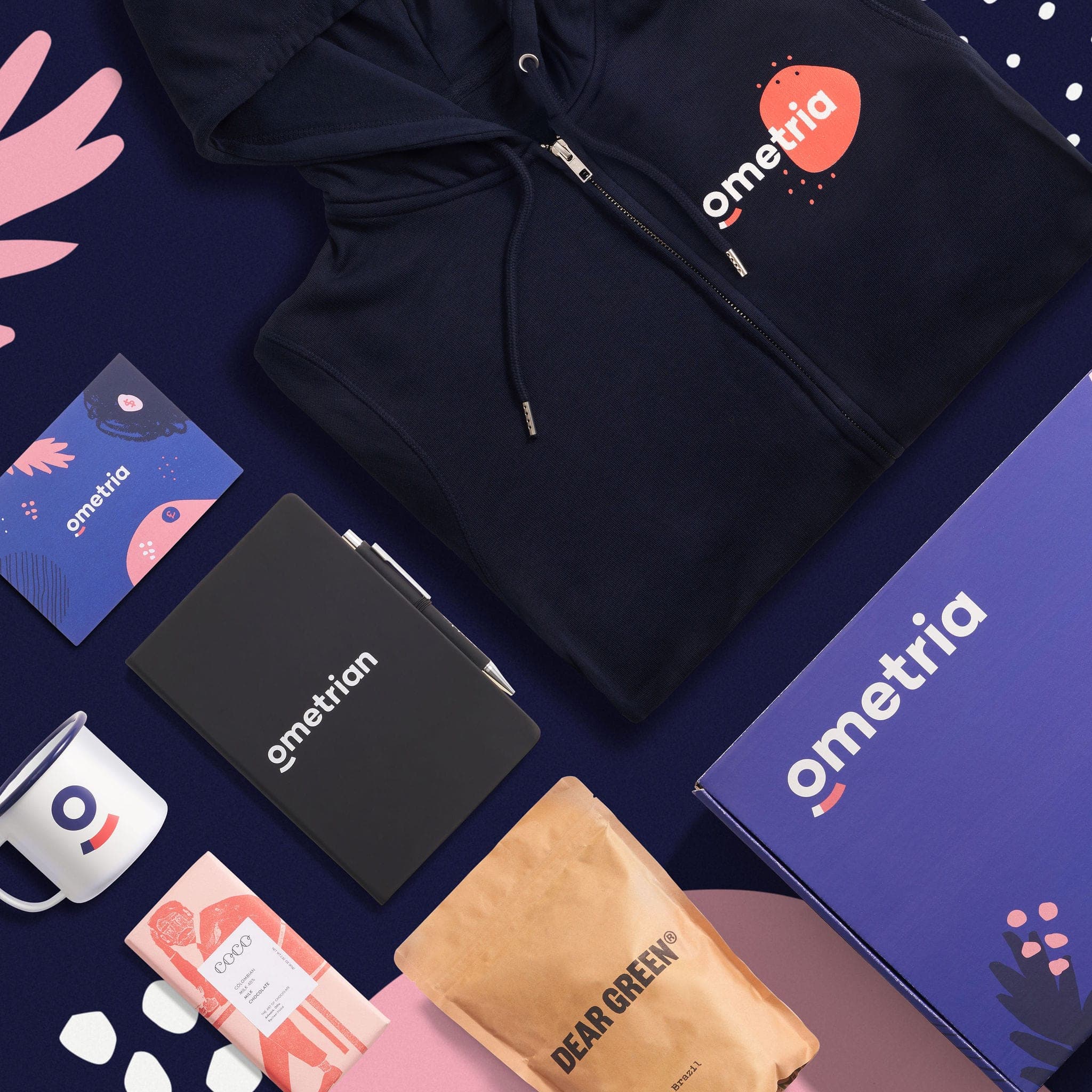 Angled view of the Ometria kit: navy hoodie, purple box, notebook, enamel mug, chocolate and Dear Green coffee