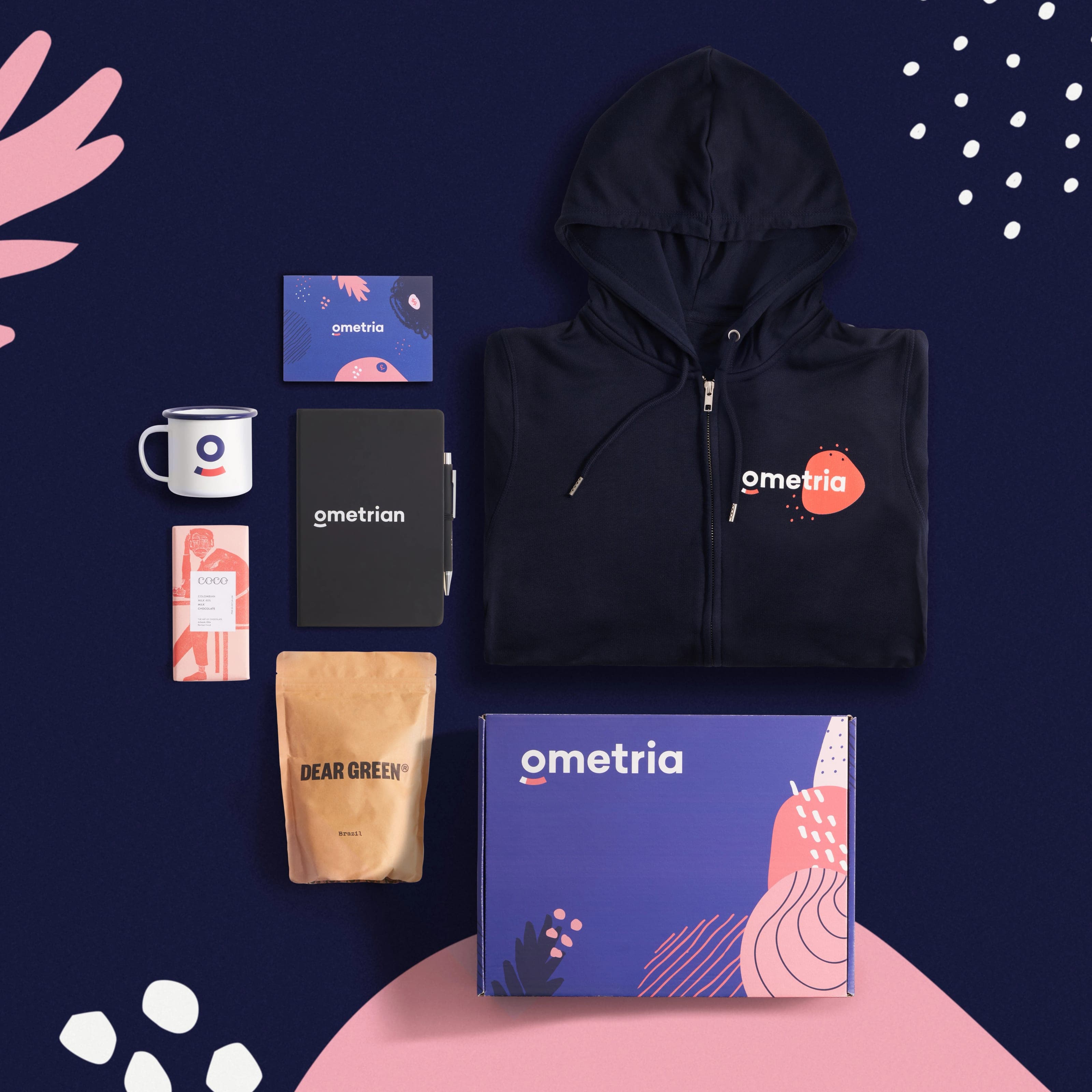 Overhead of the Ometria kit on a navy graphic with pink shapes: a navy zip hoodie, a purple mailer box, a card, a notebook and pen, a mug, coffee and chocolate