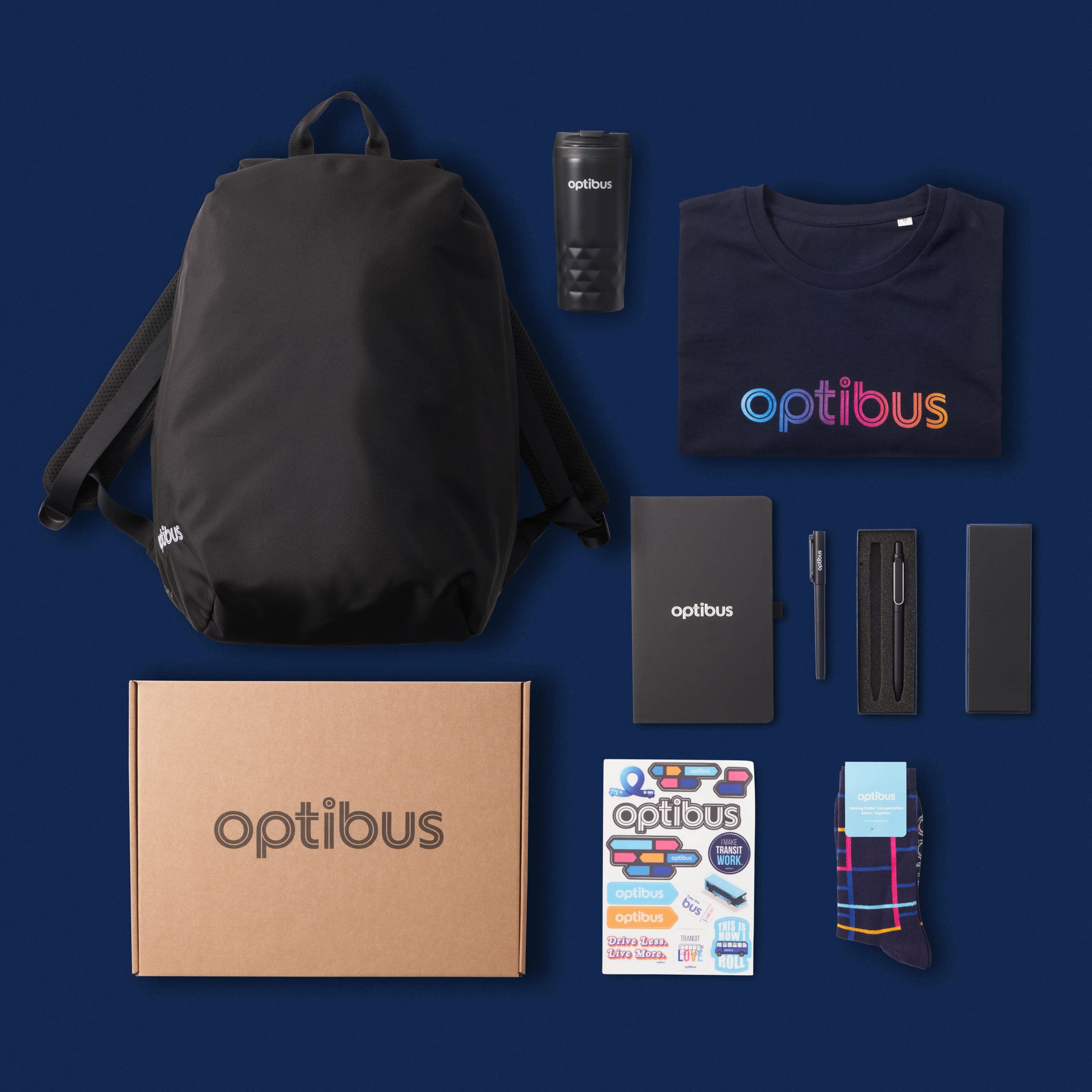 Optibus kit from above on navy: backpack, tumbler, gradient-logo T-shirt, notebook, pens, kraft box, stickers and socks