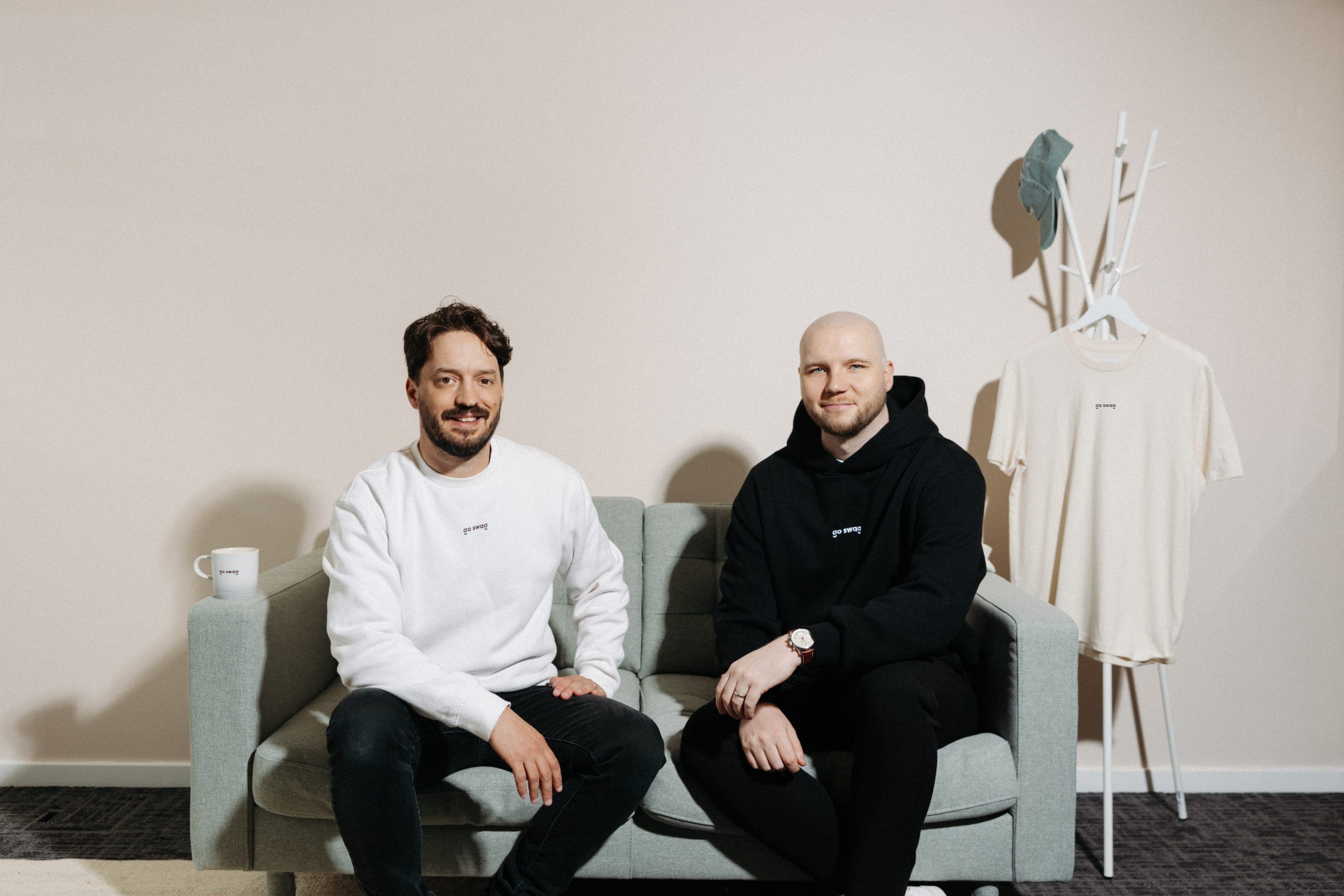 Go Swag co-founders Ben Greenock (left) and Conor McKenna sitting side by side on a sage sofa in a Go Swag sweatshirt and hoodie, with a Go Swag T-shirt and cap on a coat stand beside them