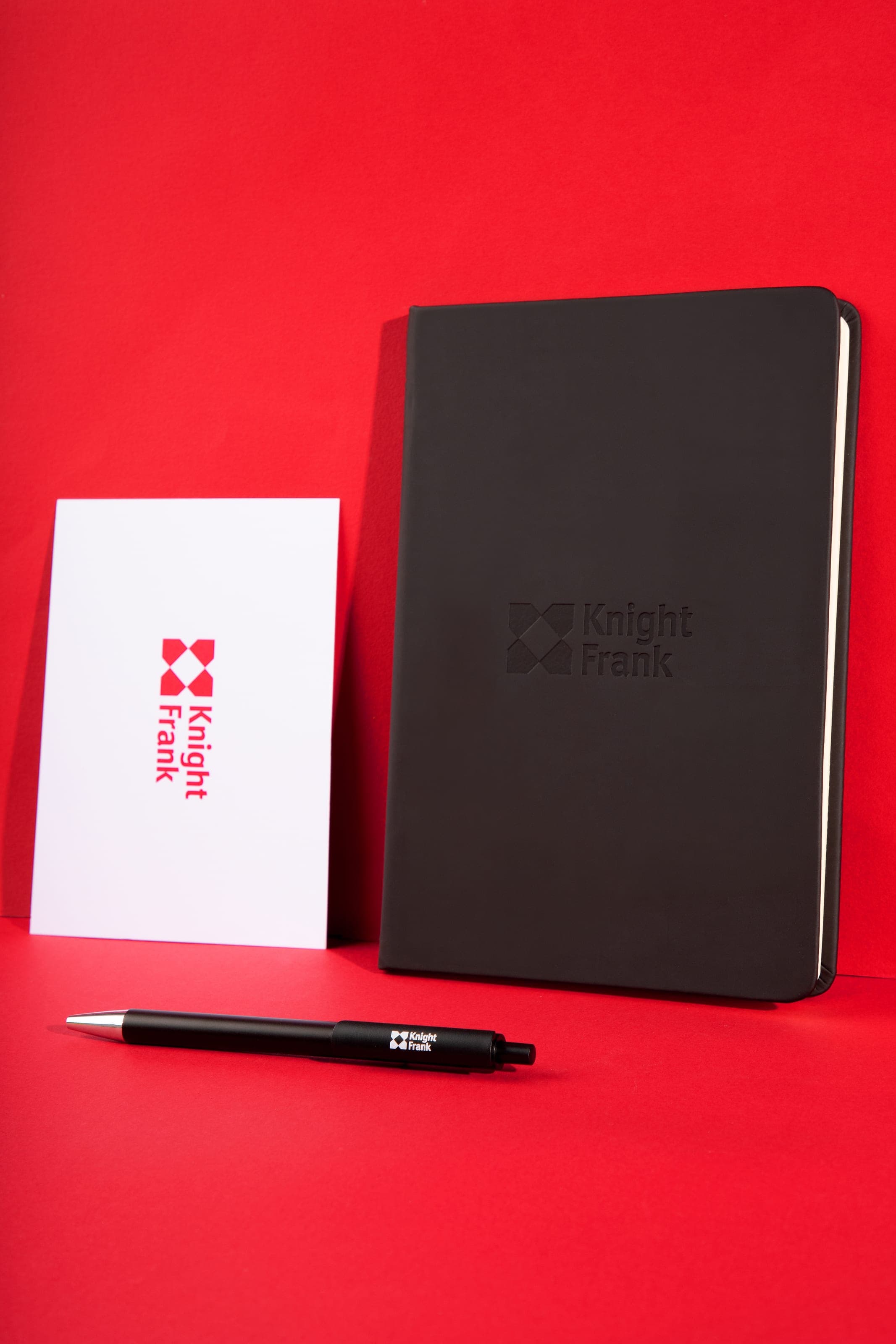 A debossed black Knight Frank notebook beside a white Knight Frank card and a black pen on a red set