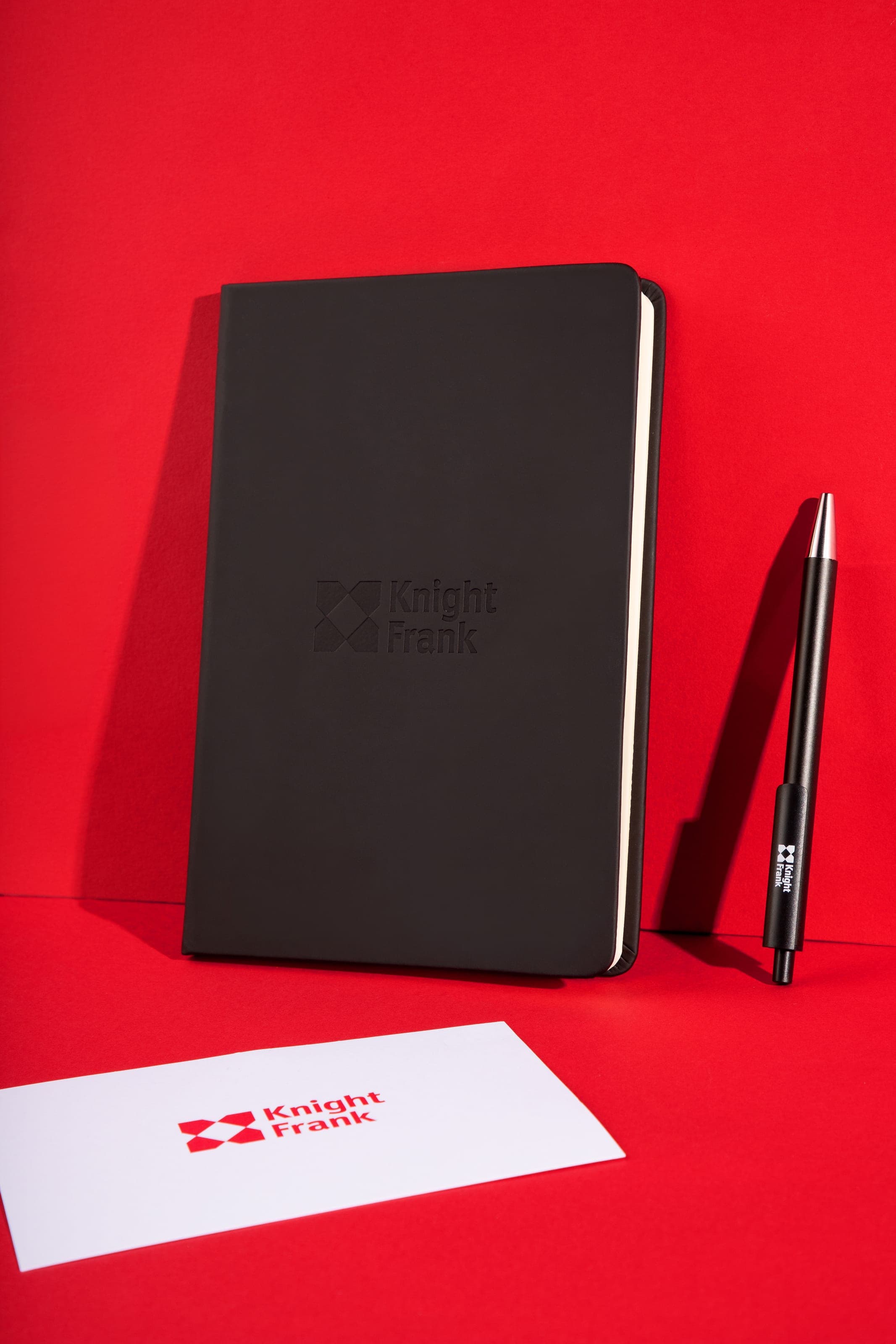 Black notebook debossed with the Knight Frank logo standing on red, with a pen leaning beside it and a white logo card in front