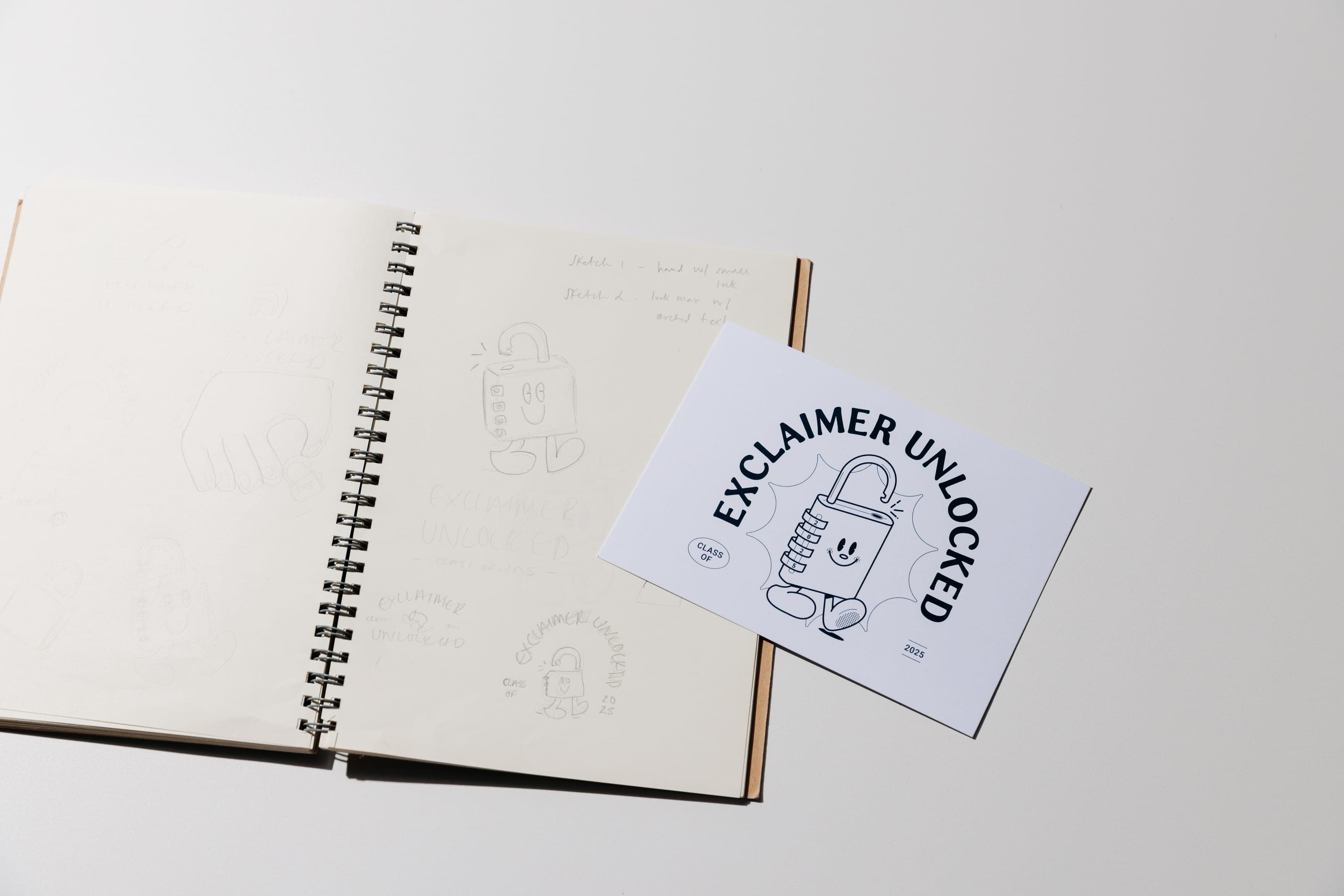 An open spiral sketchbook of padlock character drawings beside the finished Exclaimer Unlocked printed card