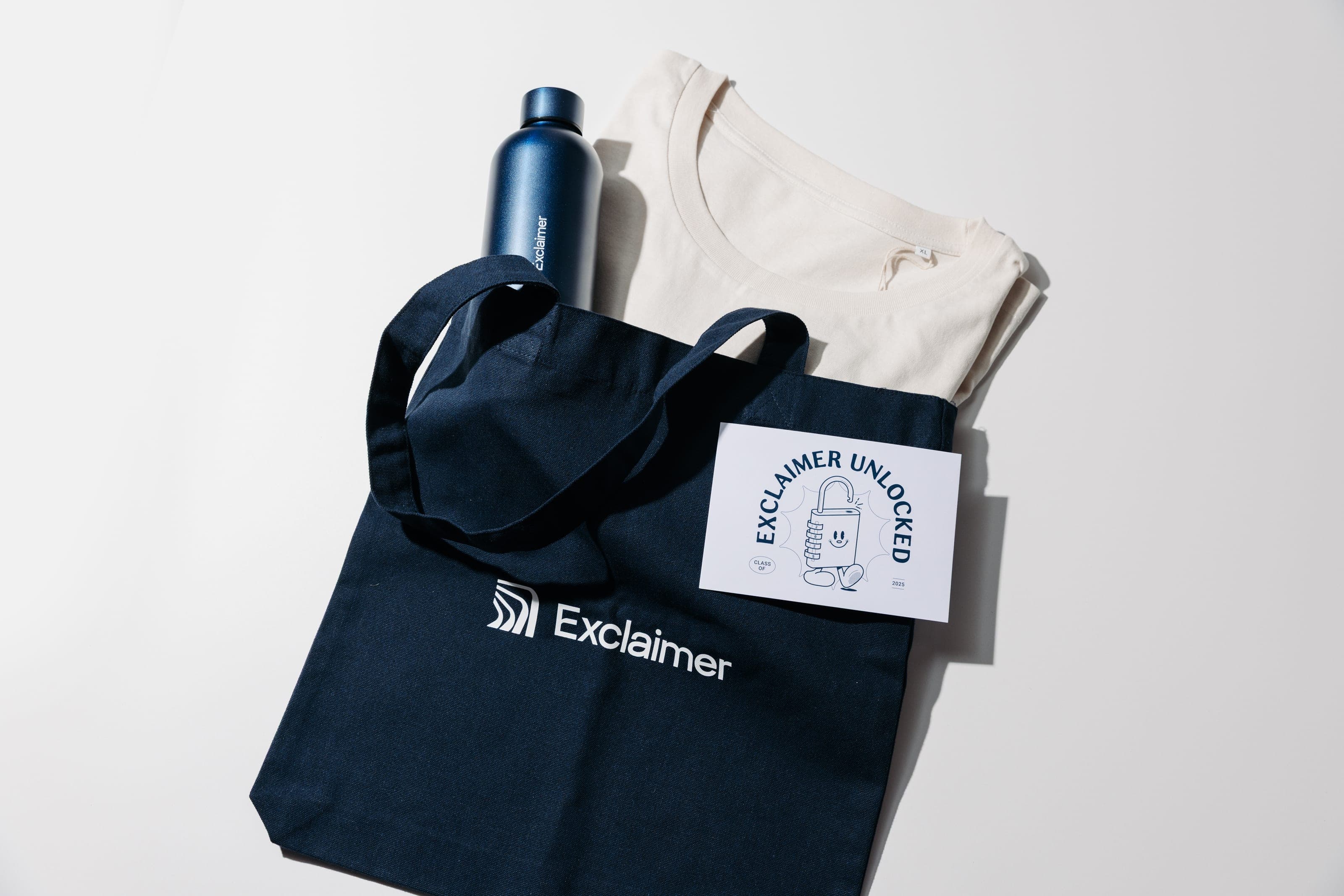Flat lay of a navy Exclaimer tote, a navy Exclaimer bottle, a folded cream T-shirt and an Exclaimer Unlocked card on white