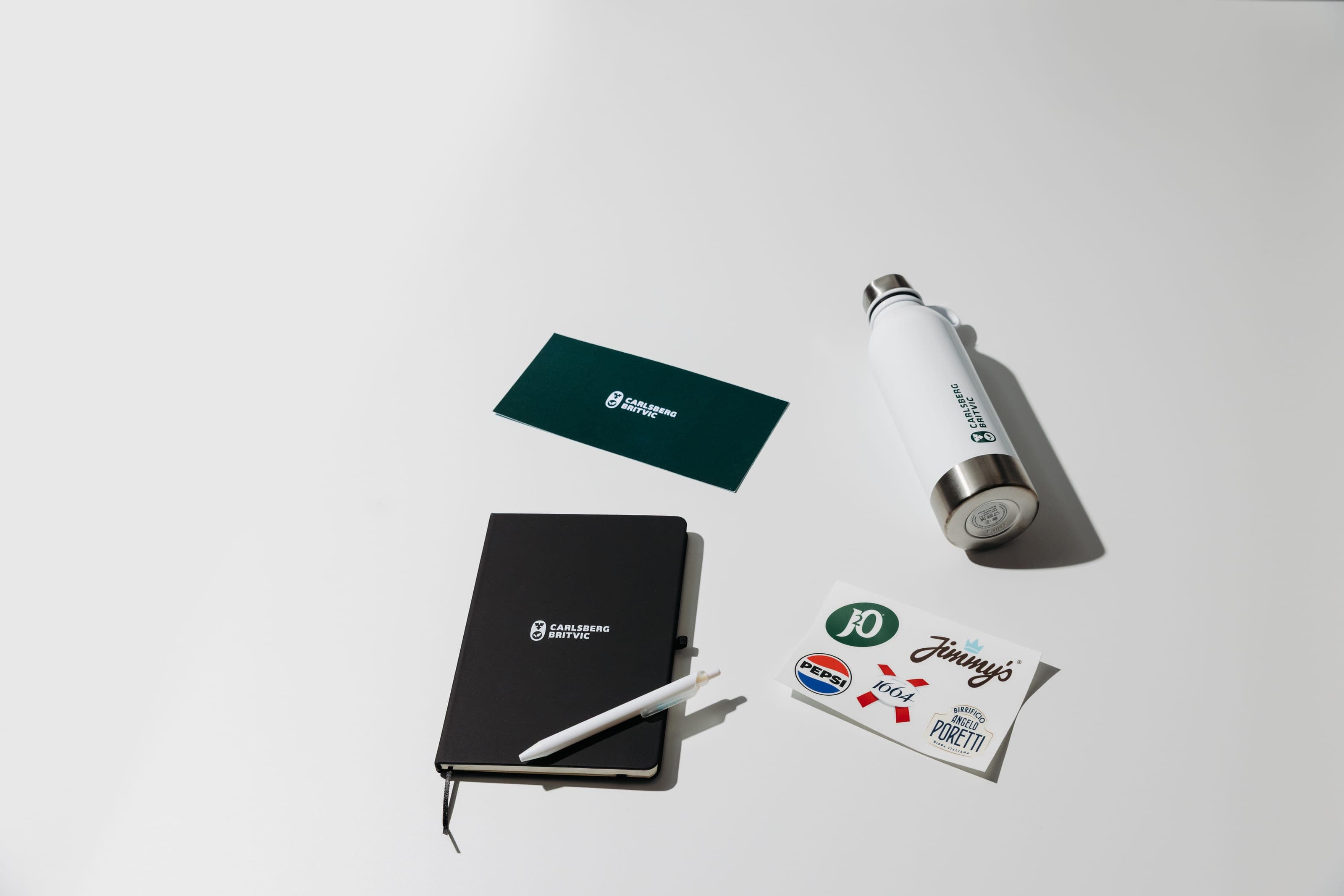 Flat lay of a green Carlsberg Britvic card, a white bottle, a black notebook with a white pen and a sheet of drinks-brand stickers on white