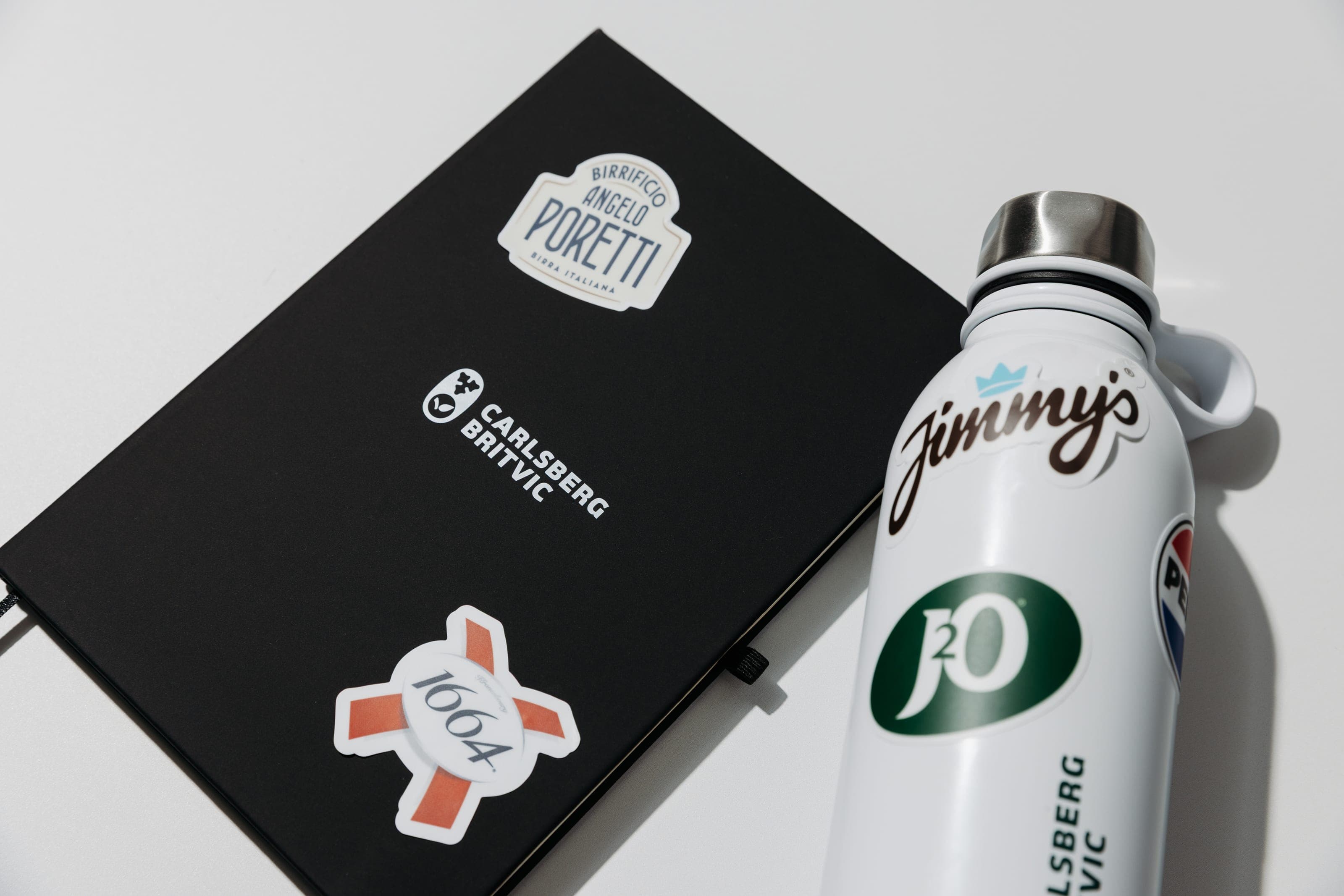 Black Carlsberg Britvic notebook and white bottle covered in Poretti, 1664, Jimmy's and J2O stickers