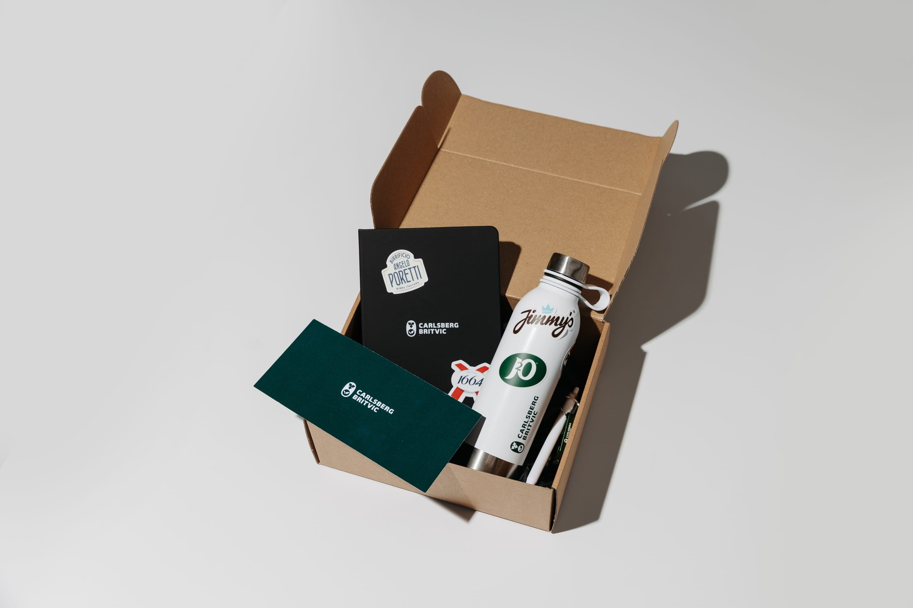 Open kraft box holding a Carlsberg Britvic notebook, white bottle, green card and pen